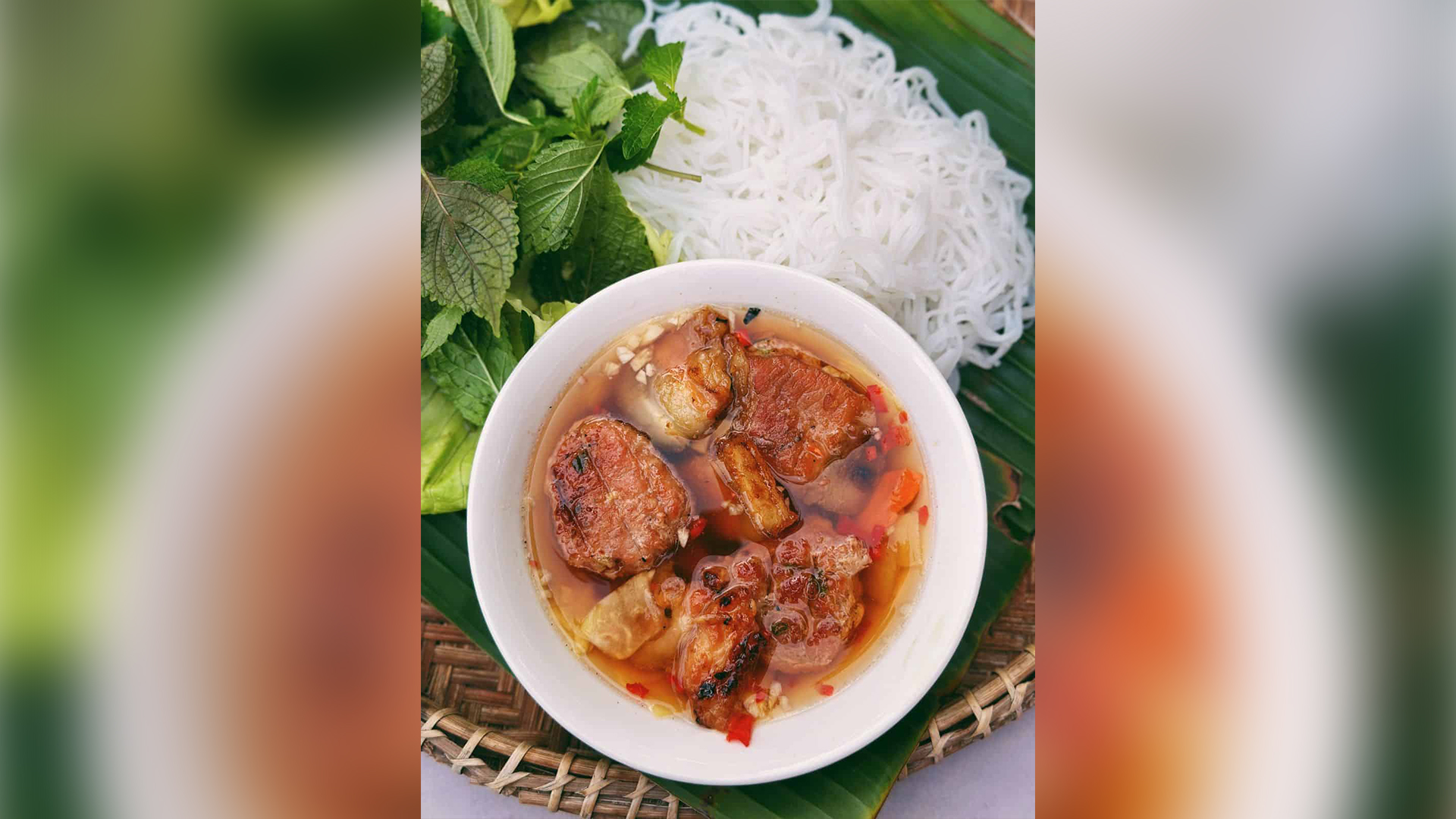 A white bowl of Vietnamese bun cha containing grilled pork patties in broth, served alongside white rice vermicelli noodles and fresh green herbs on a banana leaf.