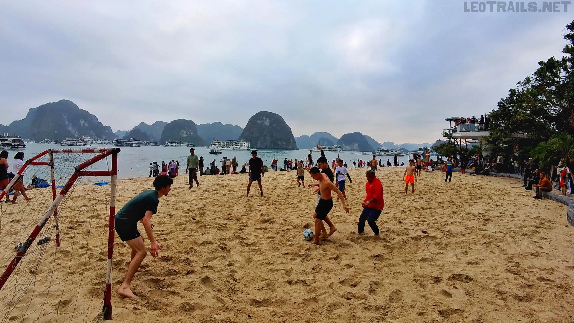 Wide-angle shot of tourists playing an energetic game of beach soccer on the sandy shores of Ti Top Island beach in Ha Long Bay, Vietnam. A red metal soccer goal sits on the left foreground, while players chase a soccer ball across the sand, with traditional cruise boats and towering limestone karsts dotting the sea under a cloudy sky.