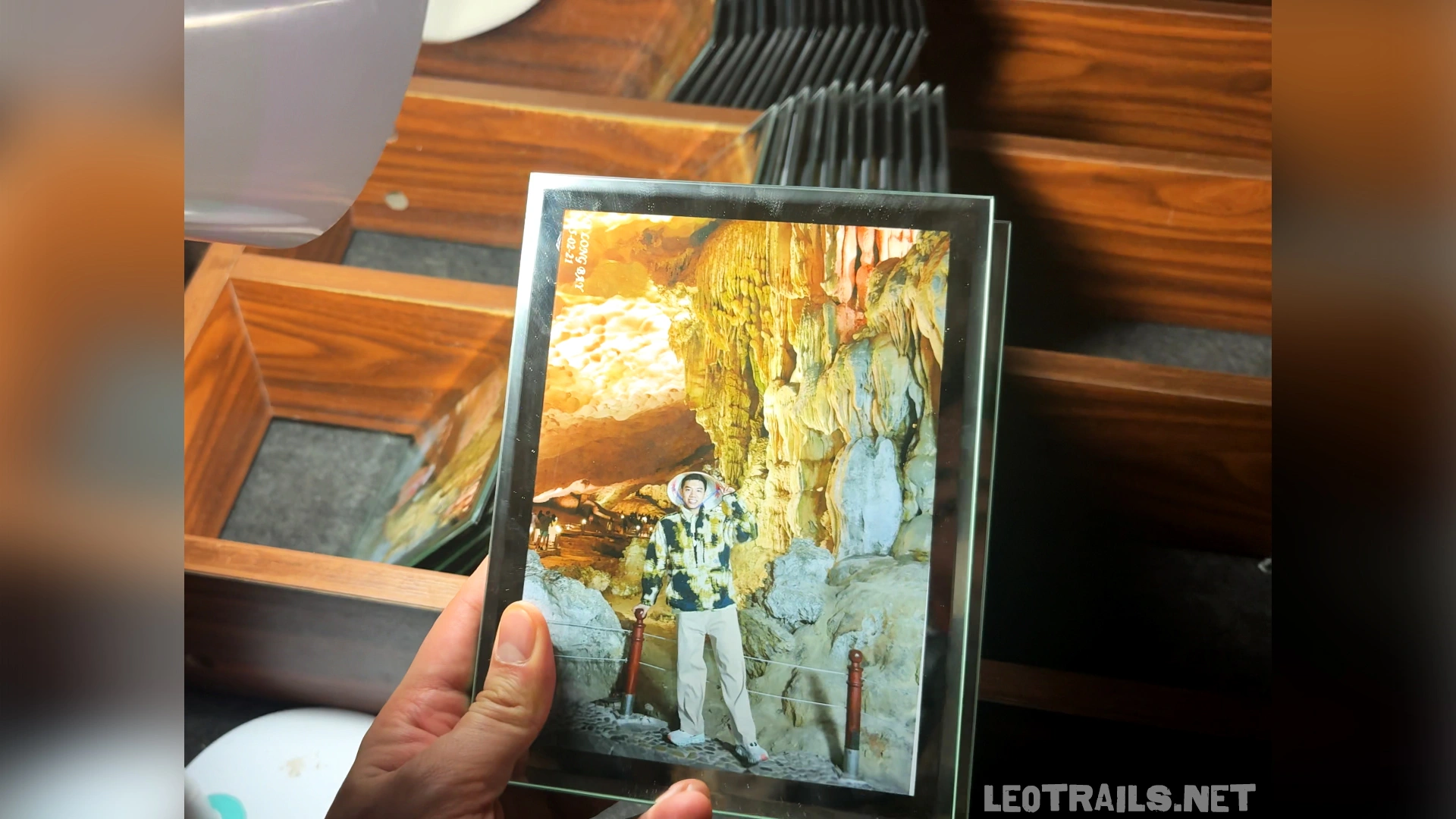 Close-up shot of a hand holding a printed souvenir photograph set inside a glass frame. The photo inside depicts a tourist posing in front of lit limestone formations inside Sung Sot Cave, with a wooden organizational rack visible in the background.