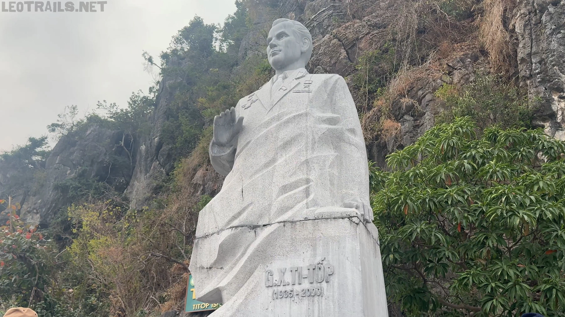 Low-angle shot of the monumental stone statue of Soviet cosmonaut Gherman Titov on Ti Top Island in Ha Long Bay, Vietnam. The gray bluestone sculpture depicts Titov in a suit with one hand raised, set against a backdrop of steep rocky cliffs and lush green trees under an overcast sky.