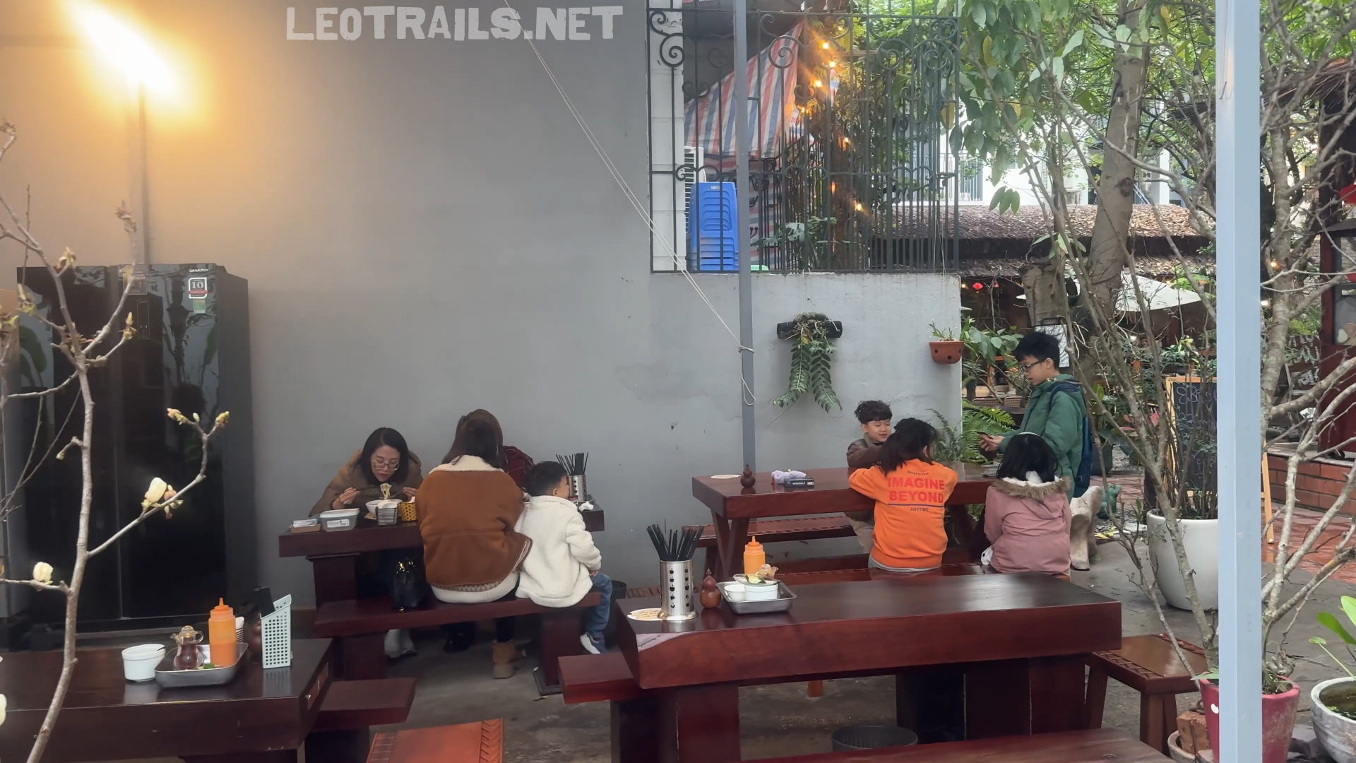 An outdoor courtyard dining area with customers seated at long wooden picnic-style tables eating noodles, surrounded by trees and greenery.