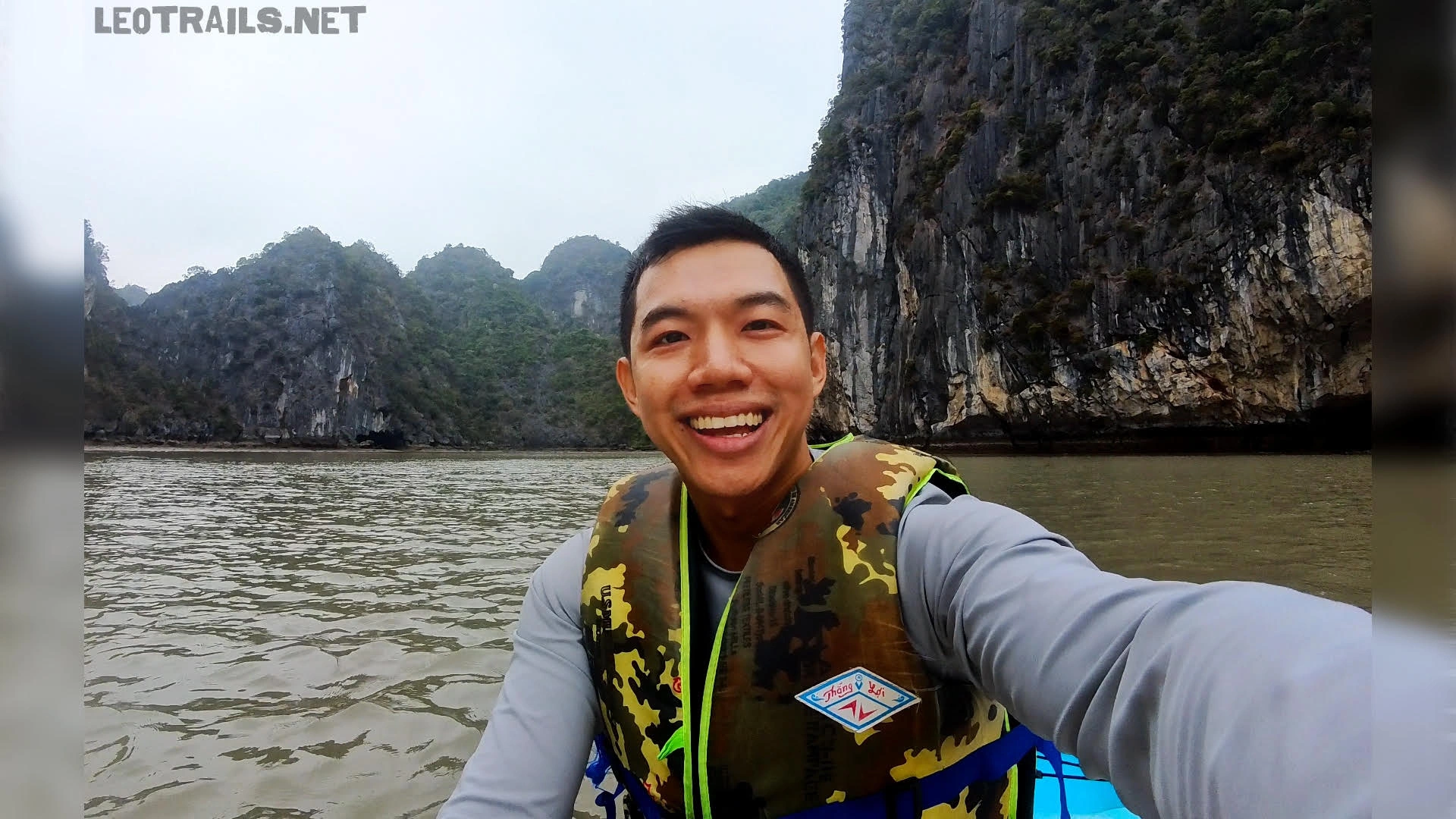 A smiling man taking a selfie while wearing a camouflage life jacket on a boat or kayak with limestone mountains in the background.