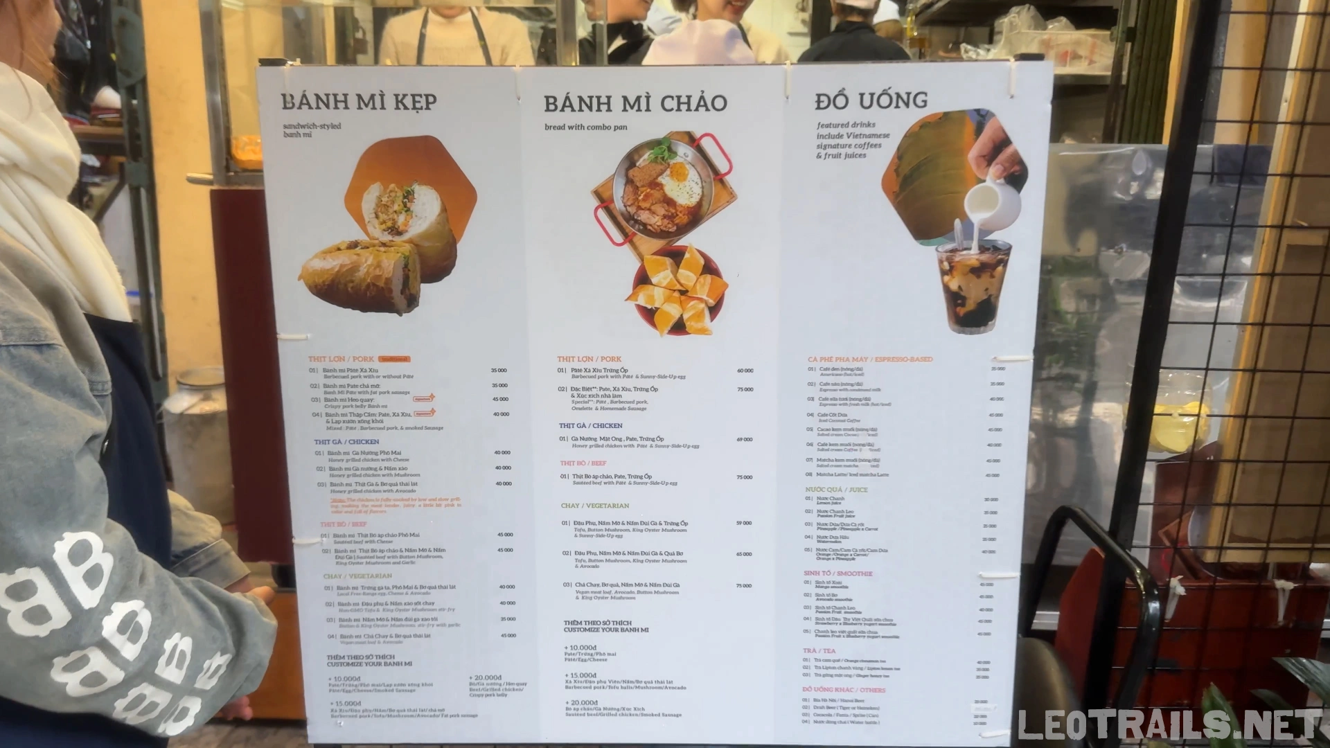A large white menu board at Banh Mi 25 displayed outdoors, split into three sections for Bánh Mì Kẹp (sandwiches), Bánh Mì Chảo (skillets), and Đồ Uống (beverages) with clear English translations and prices.