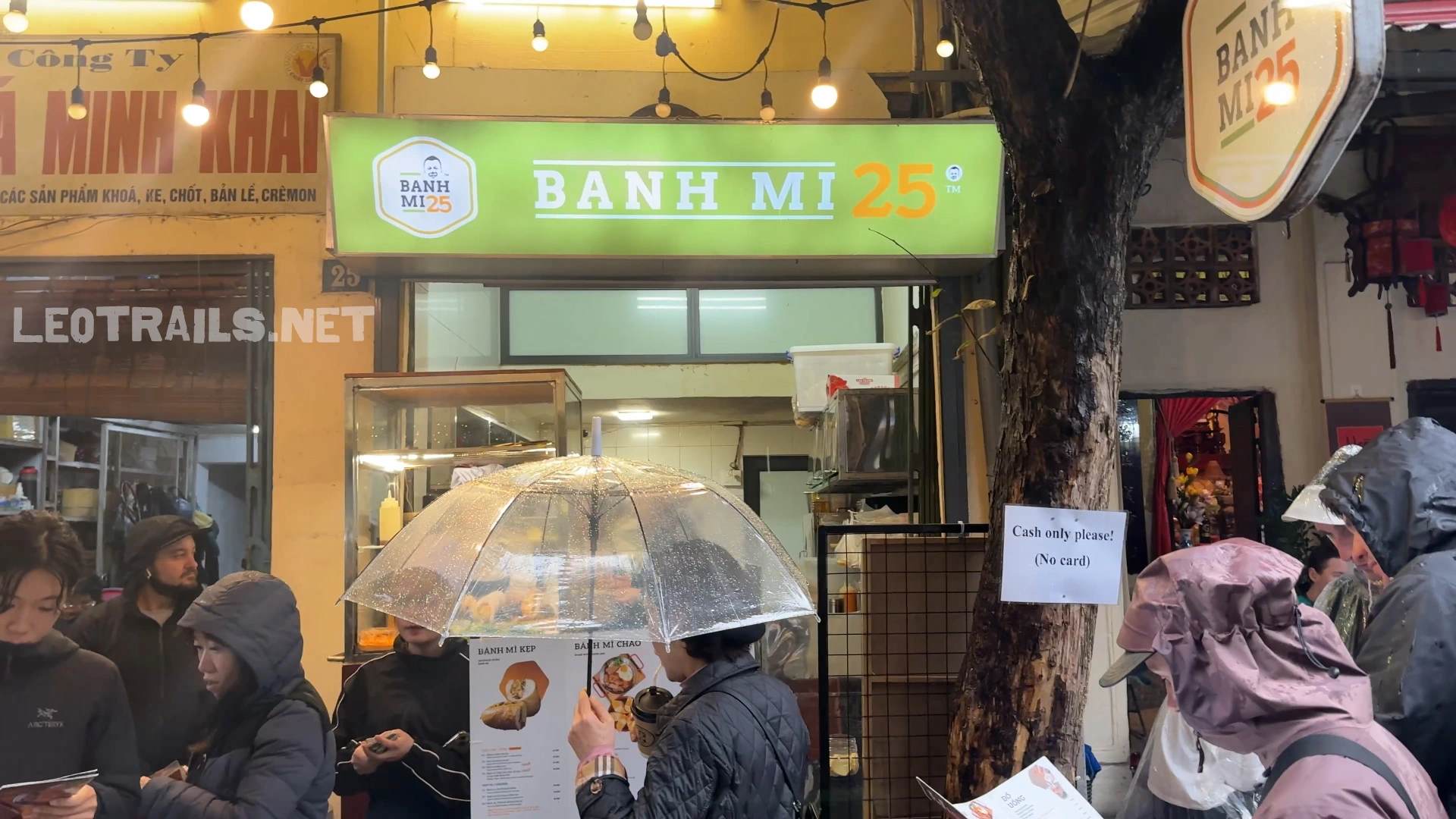 The street-facing storefront of Banh Mi 25 in Hanoi, Vietnam, showing a green illuminated sign with string lights above and customers holding umbrellas waiting in line outside.
