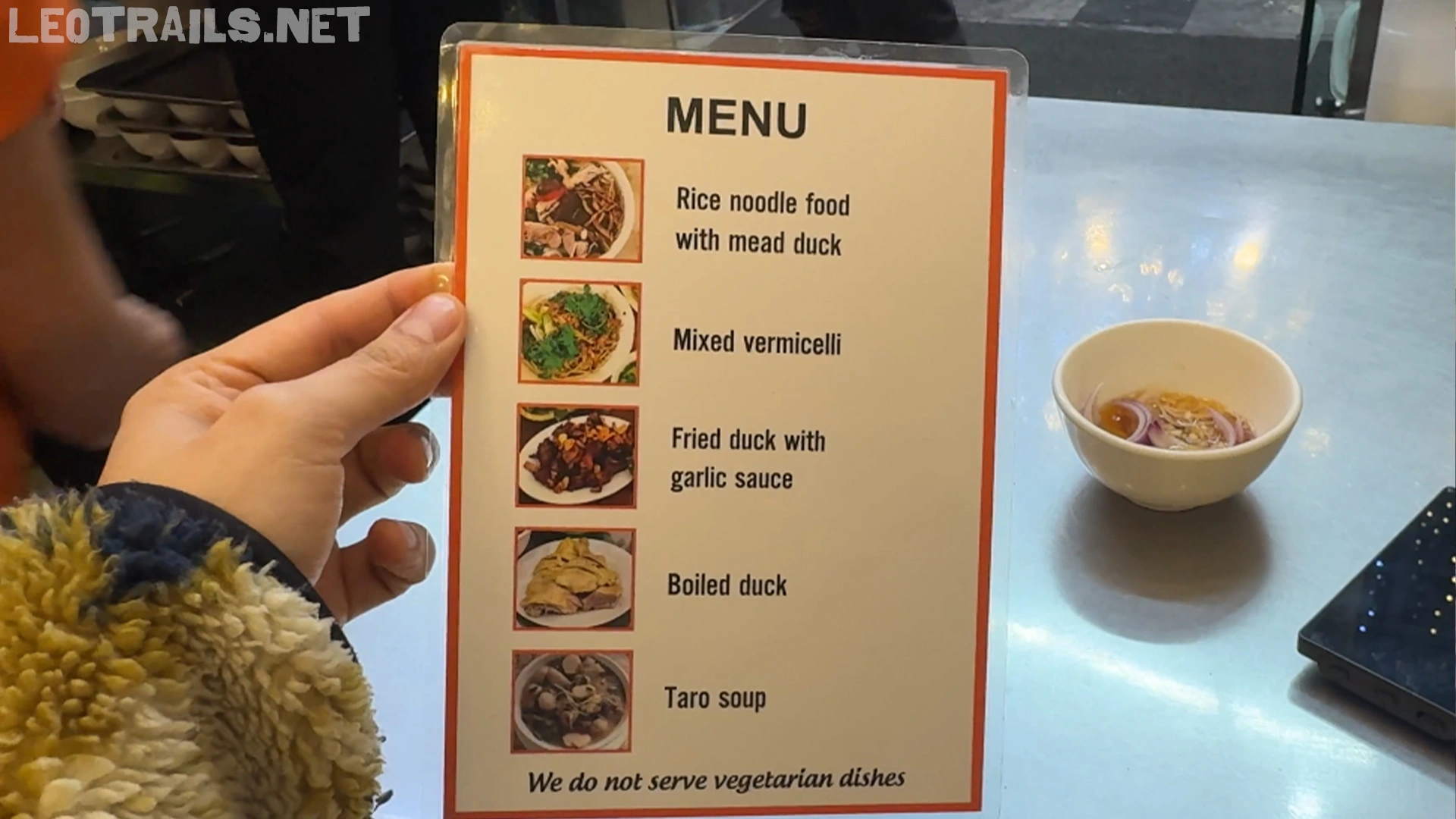 A hand holding an English menu card listing duck dishes like rice noodle food with mead duck, mixed vermicelli, and fried duck with garlic sauce, with a small text at the bottom stating "We do not serve vegetarian dishes".
