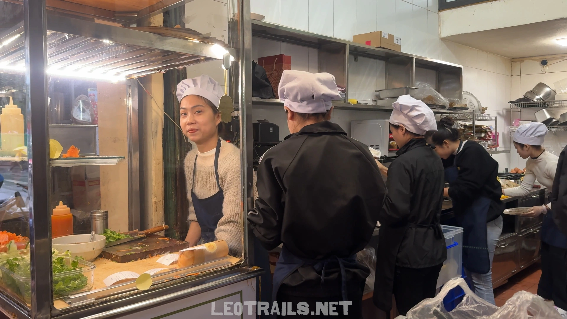 Kitchen staff wearing white chef hats and aprons assembling orders inside the busy open-front kitchen area of Banh Mi 25 in Hanoi.