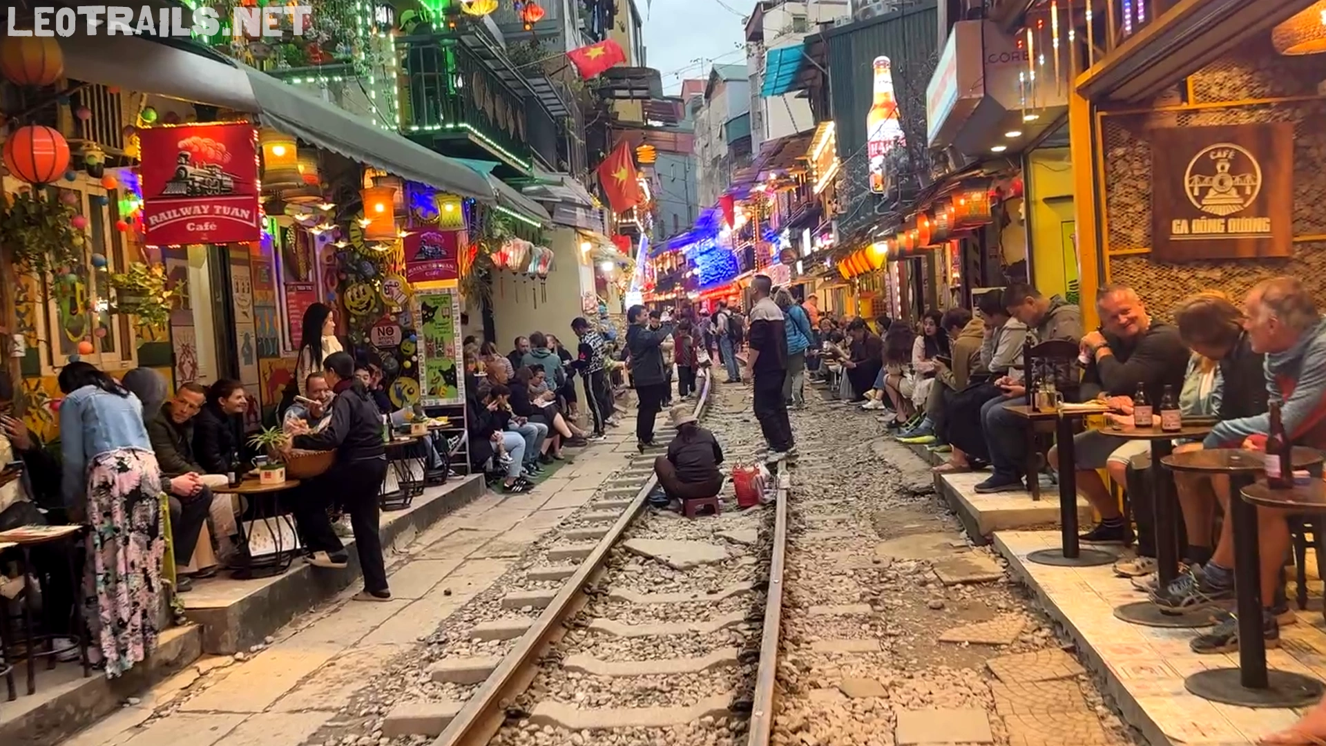 A narrow street view of Hanoi Train Street filled with tourists sitting outside vibrant, brightly lit cafes on both sides of a central railway track.