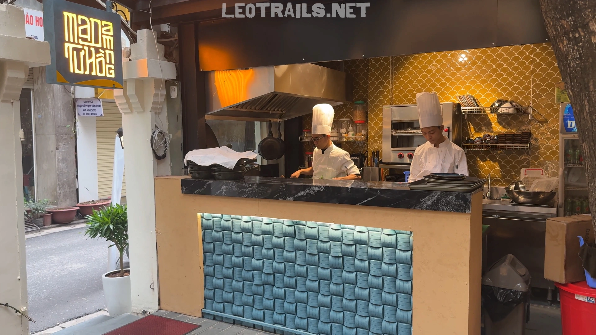 Two professional chefs in white uniforms and tall chef hats working behind an open outdoor kitchen counter at MaDam Tư Hồng restaurant in Hanoi.
