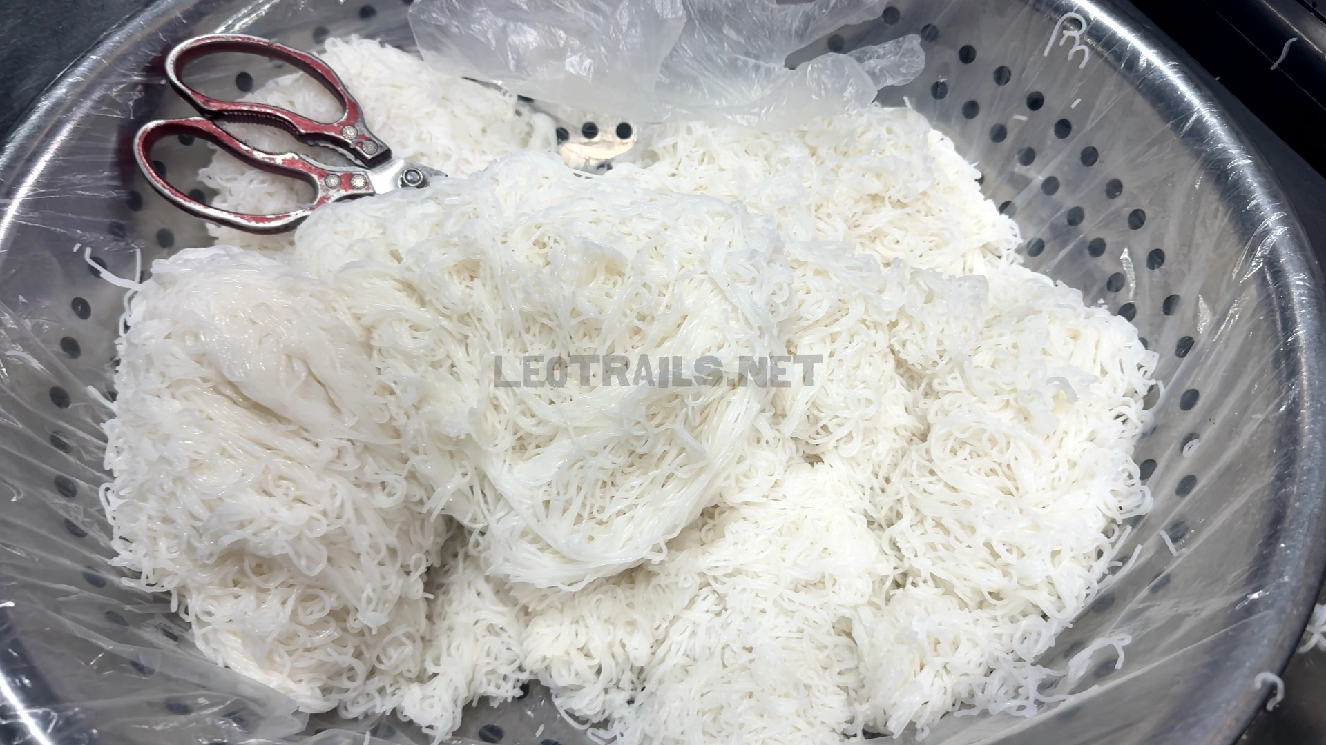 A large metal colander filled with fresh white rice vermicelli noodles (Bún) ready to be portioned and served, with a pair of kitchen scissors resting on top.