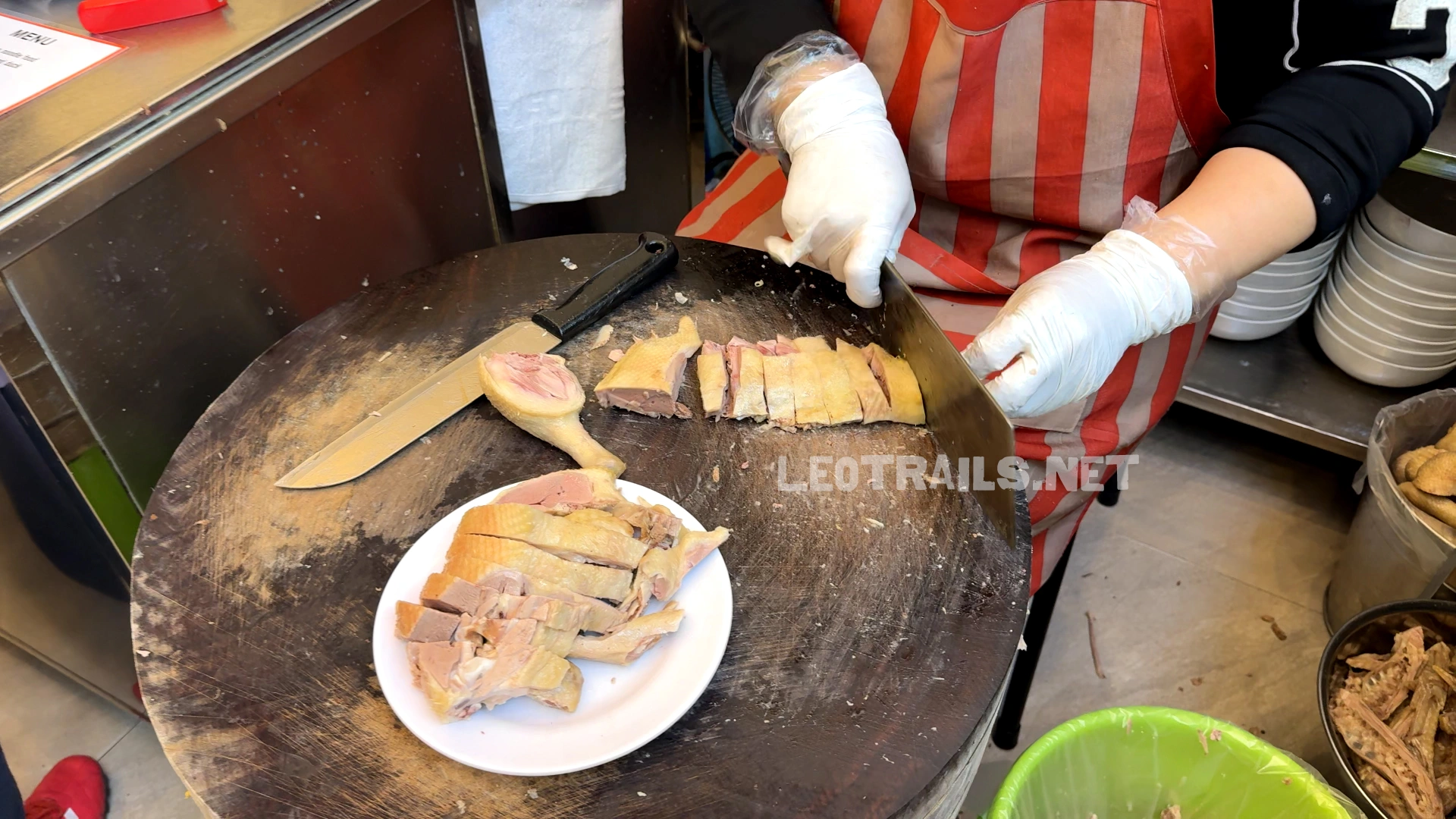 A restaurant worker wearing plastic gloves chopping succulent boiled duck meat on a large circular wooden cutting board, placing the sliced pieces onto a small white plate.