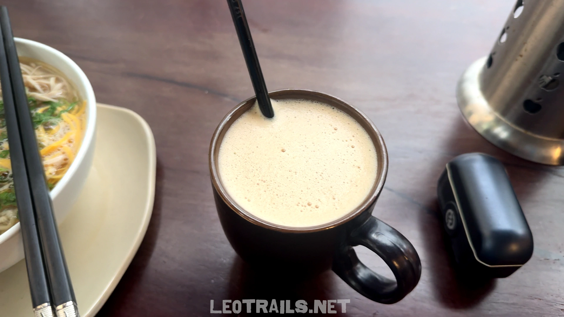 A dark brown mug filled with frothy Vietnamese coconut coffee sitting on a wooden table next to a bowl of noodle soup.