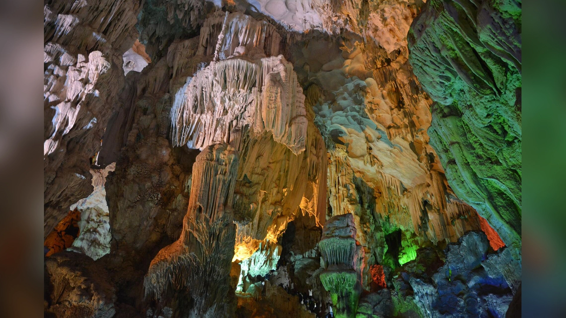 Wide-angle interior view of Dau Go Cave (Heavenly Palace Cave) on Dau Go Island in Ha Long Bay, Vietnam. The photo displays a dramatic cavern chamber filled with massive vertical limestone columns and intricate multi-layered stalactites illuminated by a creative multicolored lighting system featuring orange, green, and blue hues.