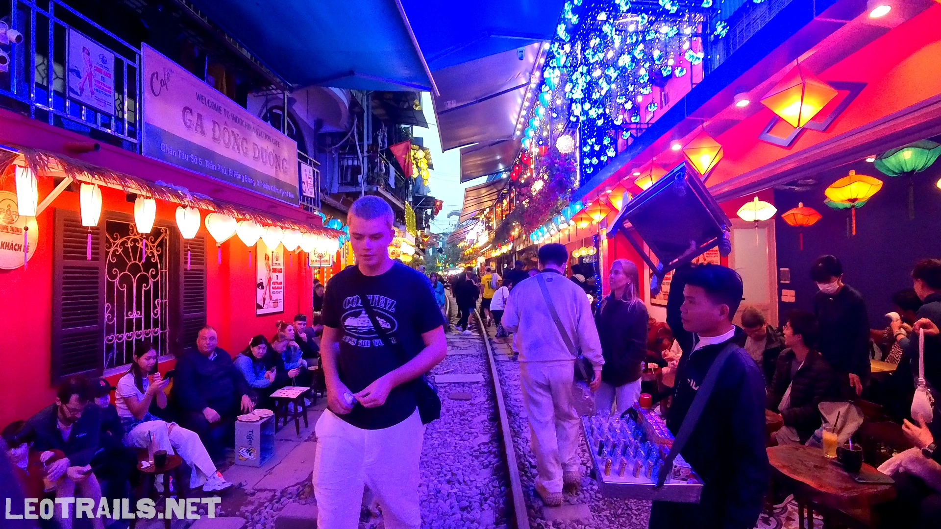 A bustling night scene at Hanoi Train Street illuminated by bright pink, blue, and orange lights, with tourists walking along the tracks past crowded cafes.