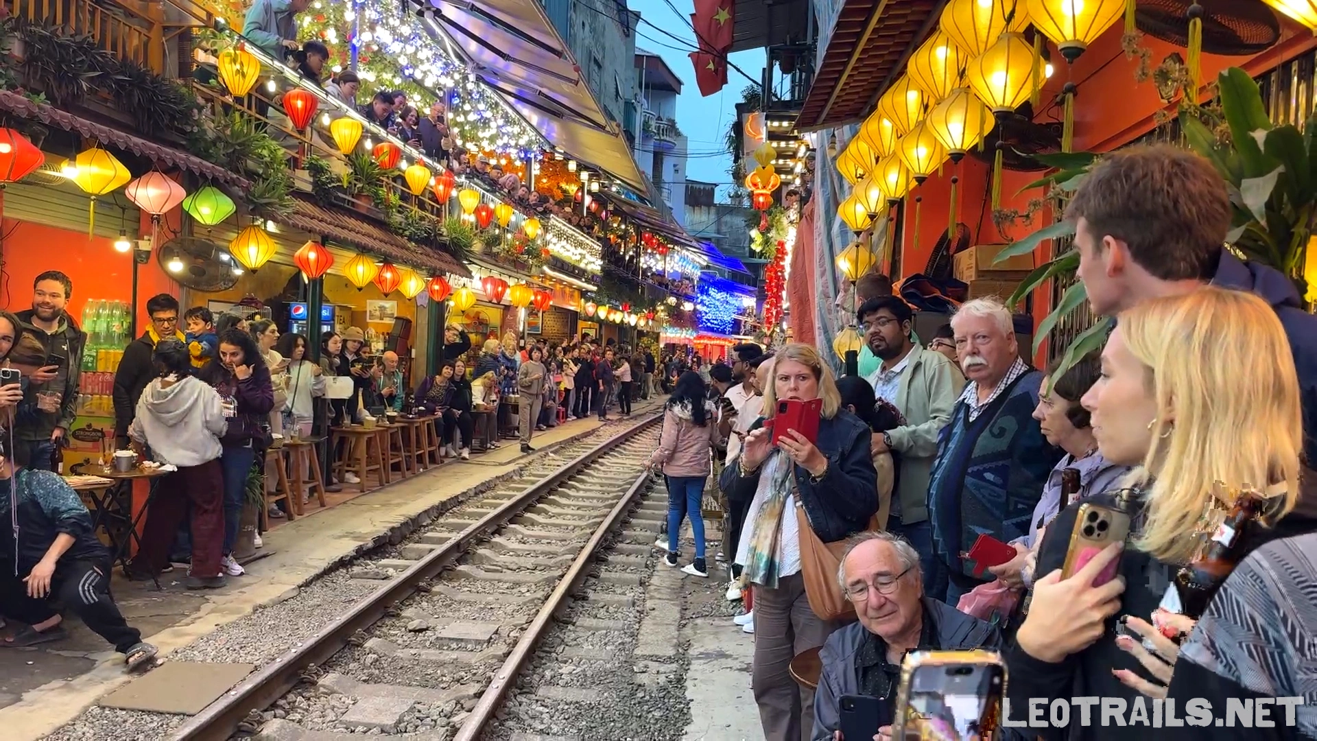 A crowded evening scene at Hanoi Train Street with tourists lining both sides of the railway tracks beneath glowing yellow and orange paper lanterns.