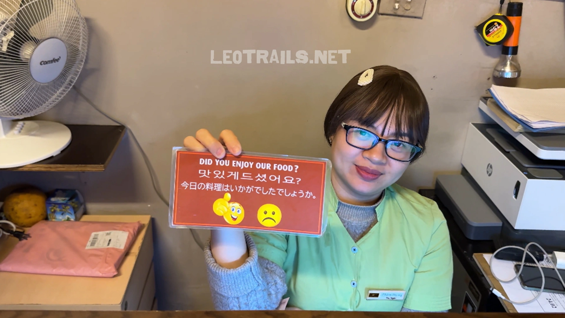 A smiling female restaurant employee wearing glasses and a green uniform holding up a red feedback sign that reads "DID YOU ENJOY OUR FOOD?" in English, Korean, and Japanese.