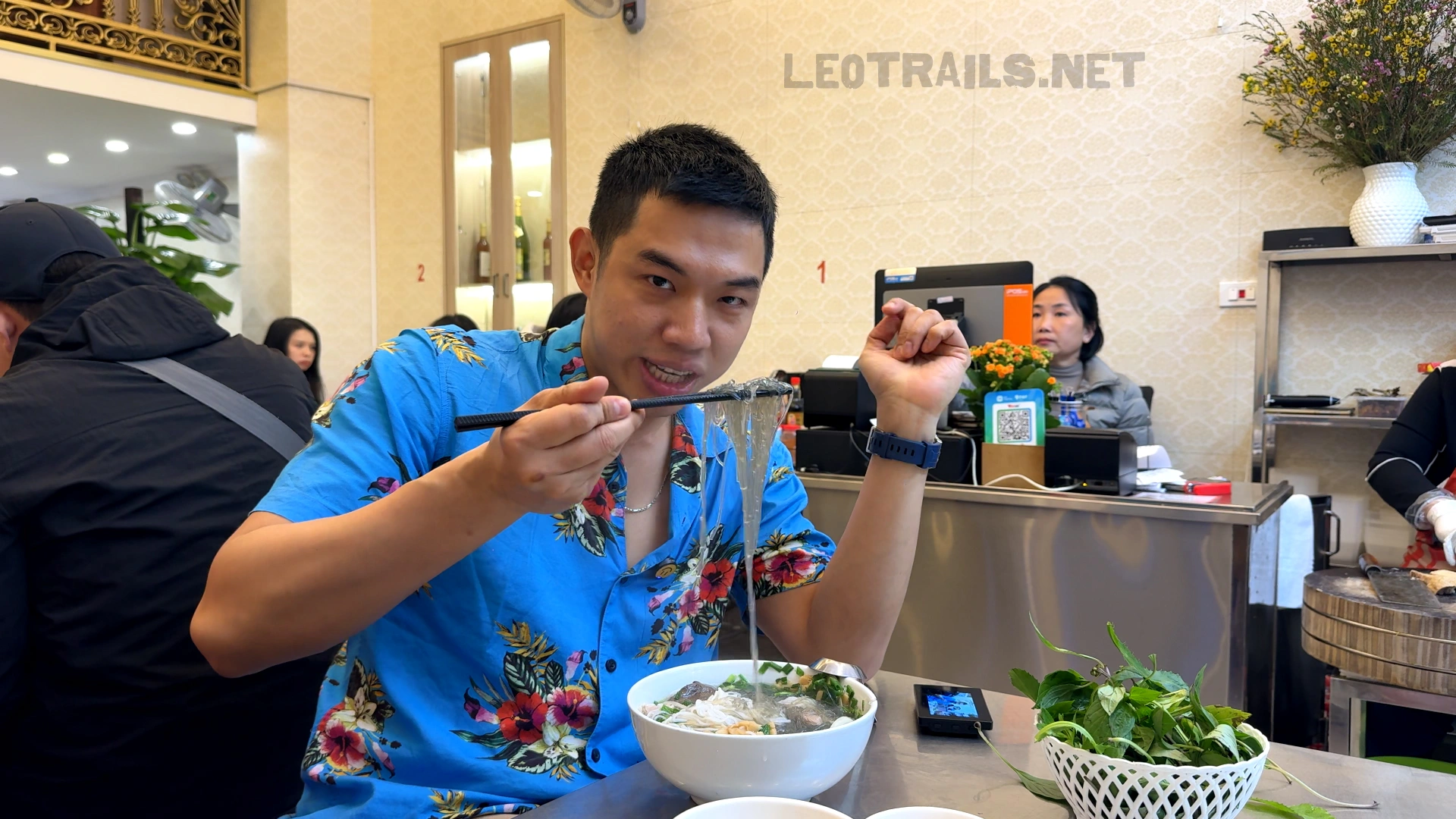 A man wearing a bright blue floral shirt lifting clear glass noodles with chopsticks from a bowl of duck noodle soup at an indoor restaurant table.