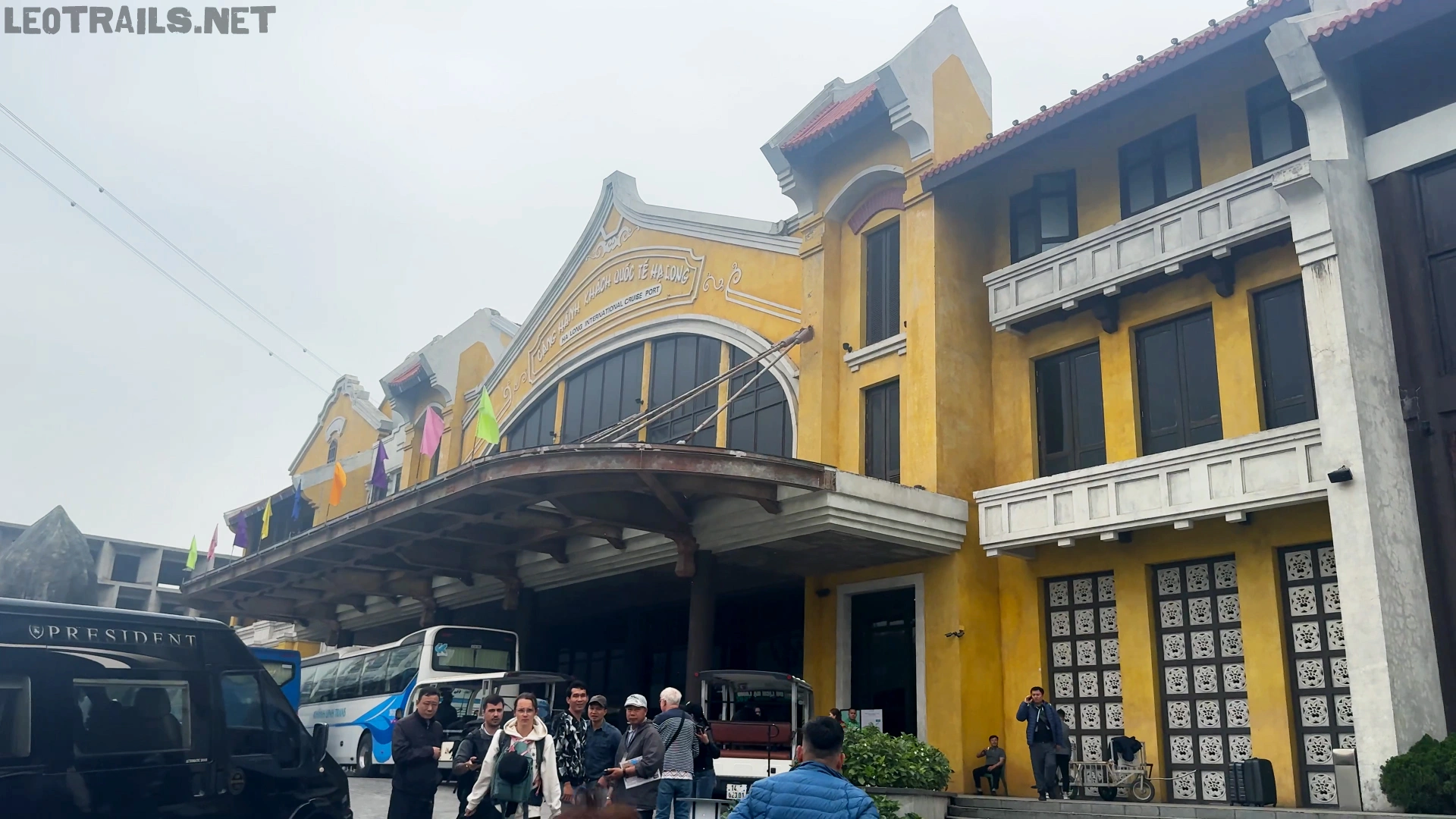 A large, colonial-style yellow port terminal building with tourists, buses, and vans gathered out front in Ha Long.