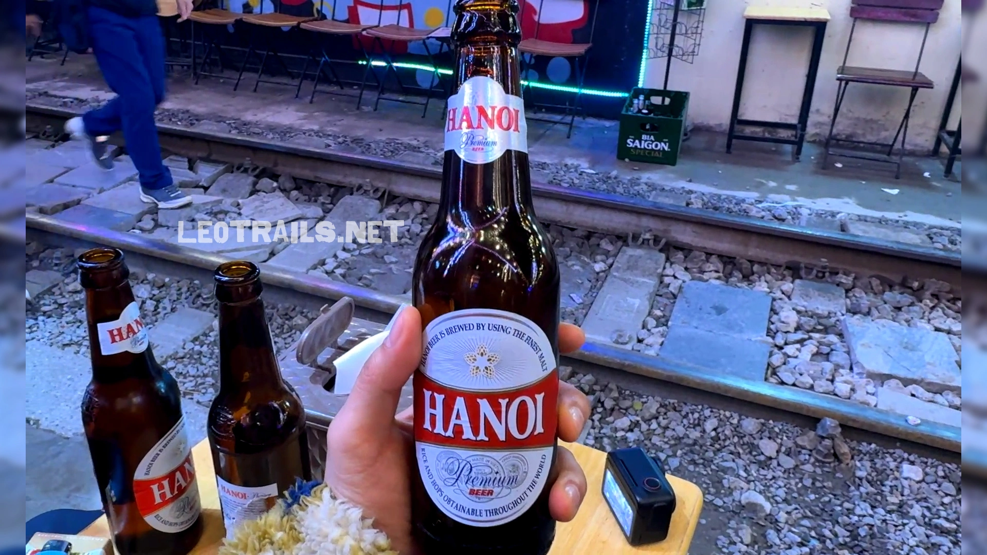 A hand holding a brown glass bottle of Hanoi Premium Beer with a red and silver label at a trackside table along Hanoi Train Street.