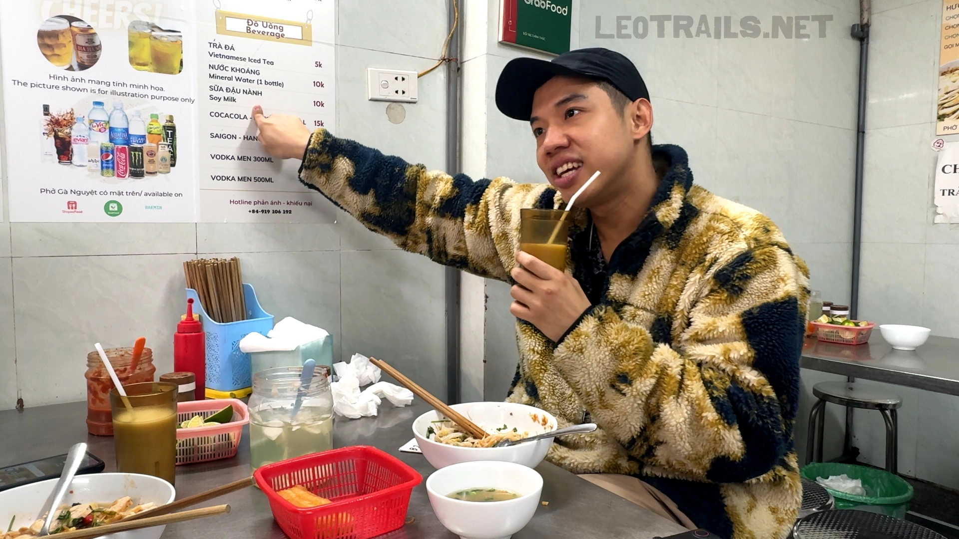 A man in a patterned fleece jacket and black cap pointing to the beverage section on the wall menu board at Phở Gà Nguyệt while holding a glass with a straw.