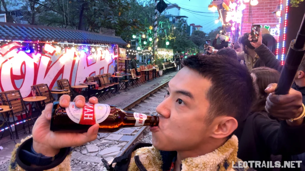 A side profile of a man drinking from a bottle of Hanoi Beer at an outdoor cafe right next to the railway tracks at Hanoi Train Street.