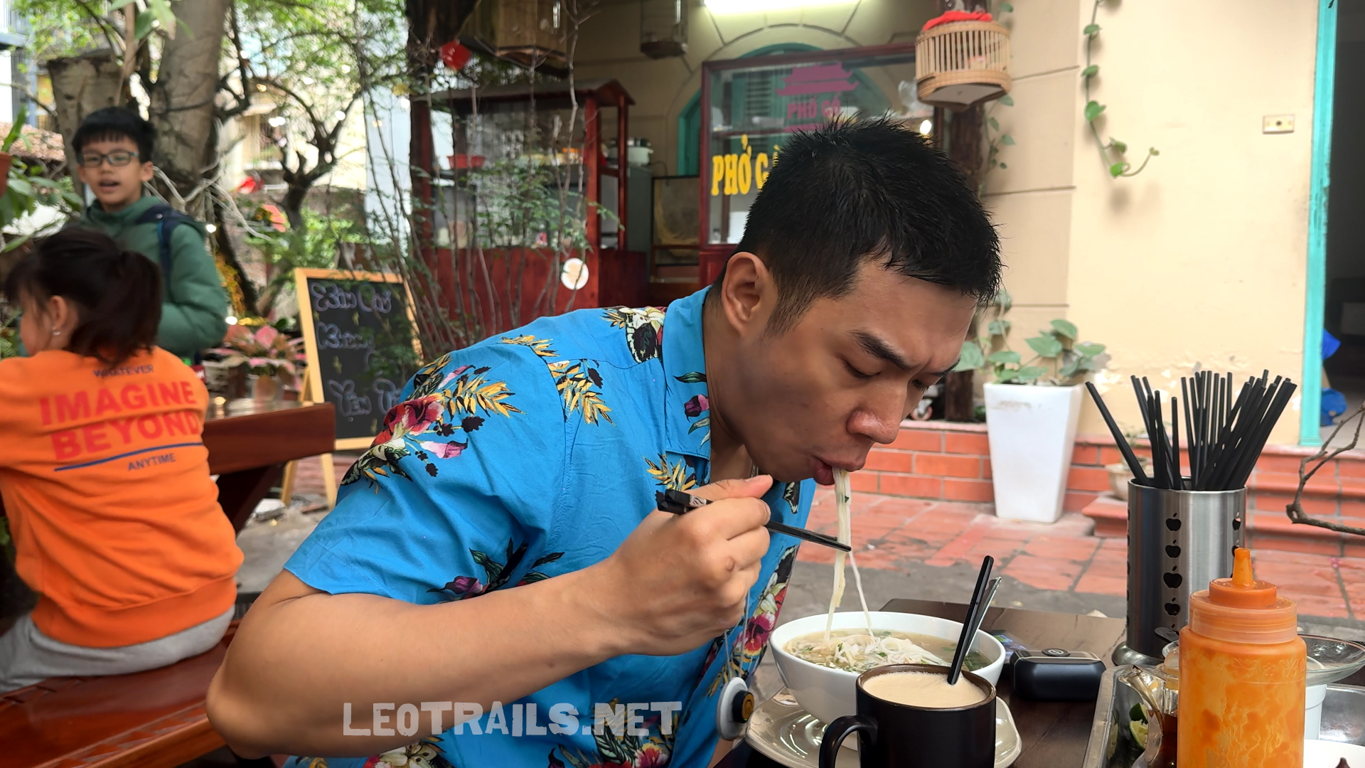 A man wearing a bright blue floral shirt eating bún thang noodle soup with chopsticks at an outdoor restaurant table in Hanoi.