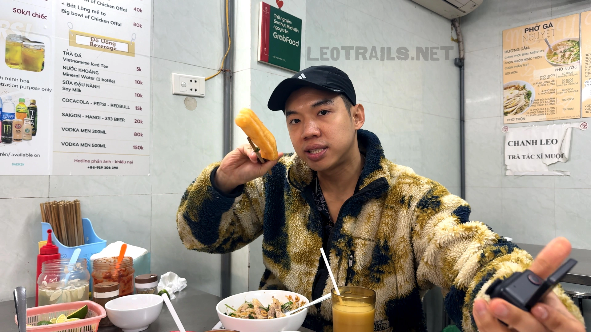 A man wearing a patterned fleece jacket and black cap holding up a crispy fried dough stick (quẩy) with chopsticks at Phở Gà Nguyệt restaurant.