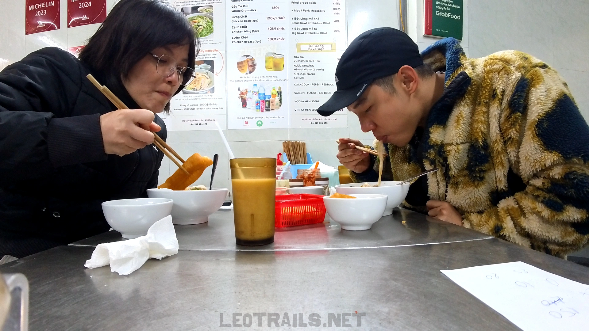 A man in a patterned fleece jacket and a woman with glasses eating chicken pho with chopsticks at a stainless steel restaurant table, with the Phở Gà Nguyệt menu board visible in the background.