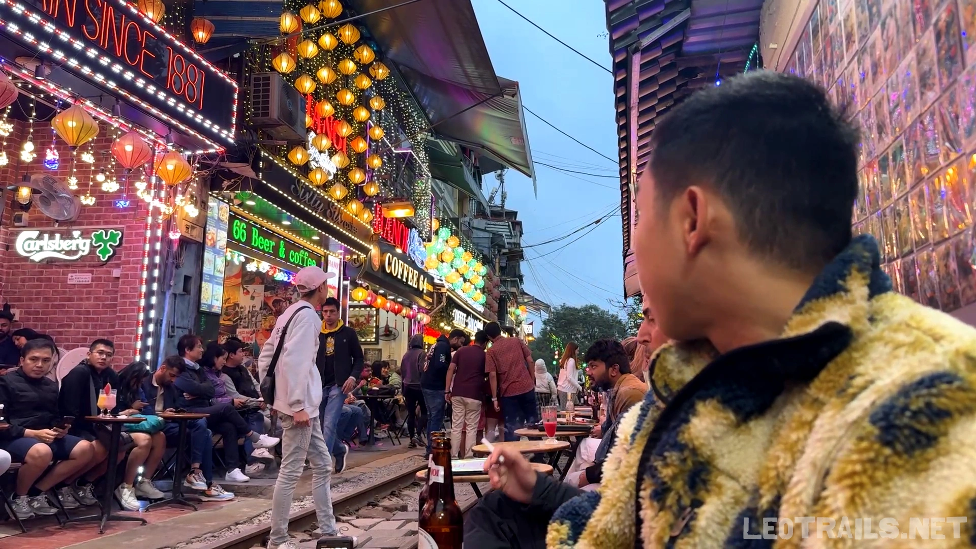 A side profile of a man in a textured jacket sitting at a trackside table, looking out over the crowded, lantern-lit railway path at Hanoi Train Street.