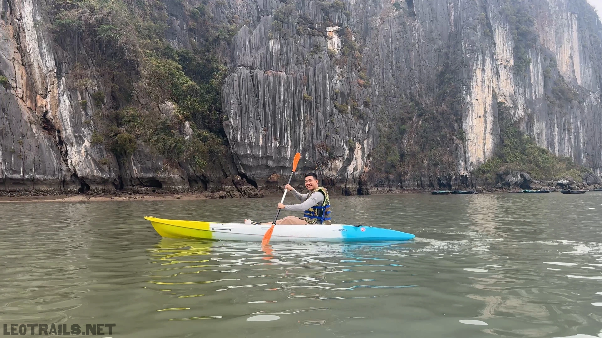 I am smiling and holding a paddle while kayaking in a yellow, white, and blue kayak on the water, with massive, steep limestone cliffs rising in the background.
