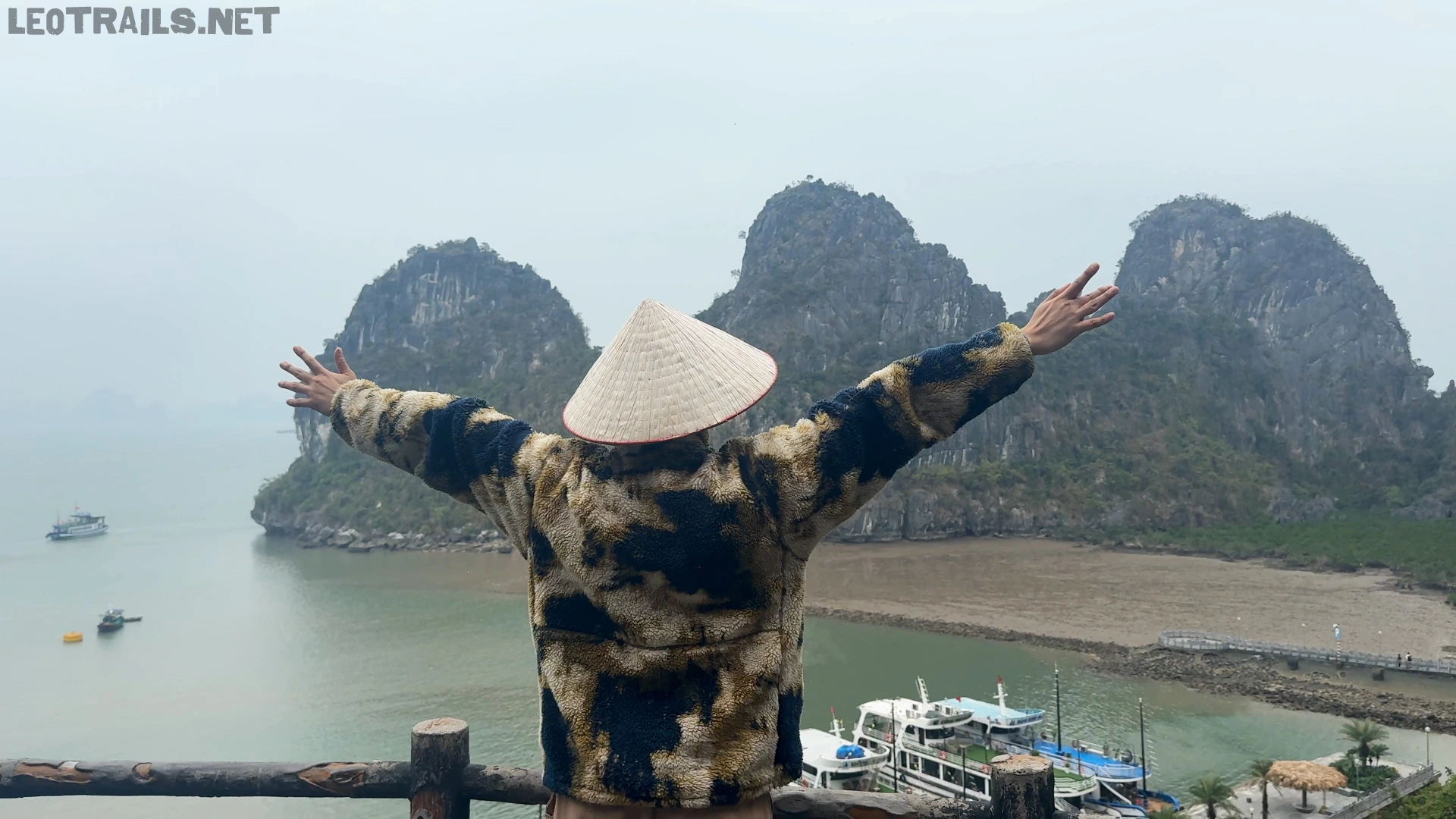 Rear view of me wearing a traditional Vietnamese conical hat (nón lá) and a camouflage jacket with arms wide open, looking out over limestone islands and boats in Ha Long Bay.