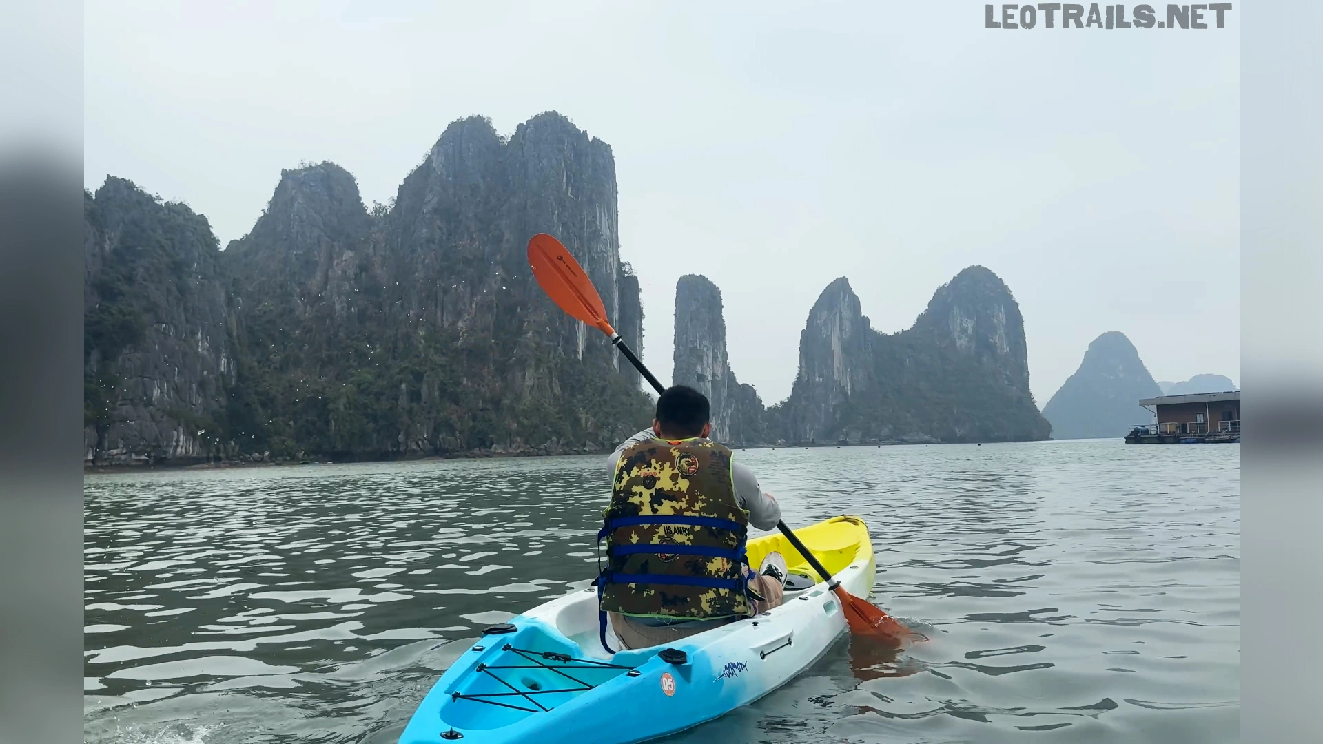 Rear view of a tourist wearing a camouflage life jacket paddling a blue and white tandem kayak across the gray waters of Ha Long Bay, Vietnam. The kayaker holds an orange paddle aloft, steering toward towering, jagged limestone karst cliffs that rise dramatically out of the misty sea under an overcast sky.