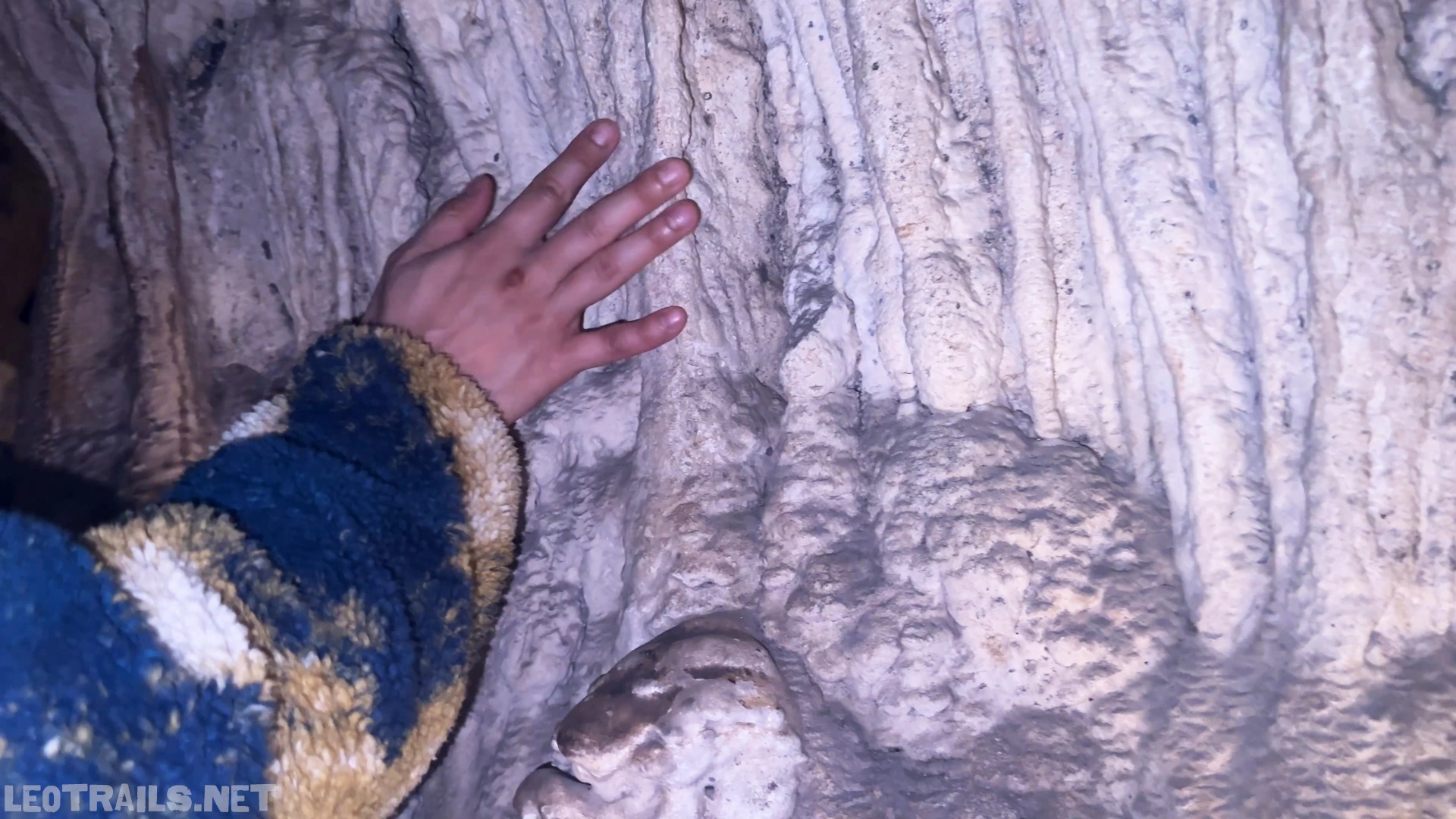 Close-up shot of a tourist's hand placing their palm against a wet, heavily ridged white limestone stalactite column inside Sung Sot Cave in Ha Long Bay, Vietnam, with the sleeve of a blue and beige fleece jacket visible.