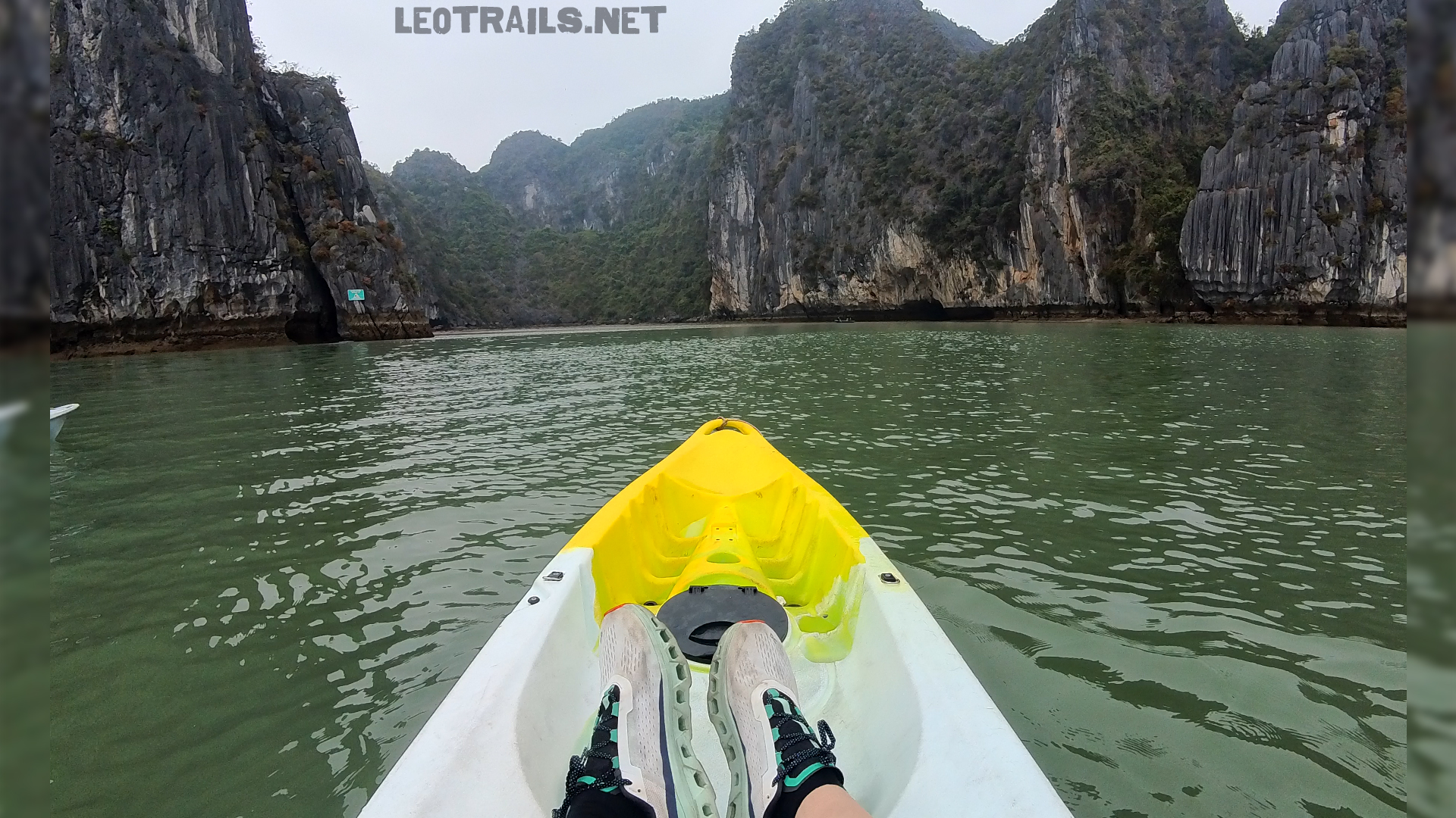 First-person perspective looking out from a white and yellow kayak, showing a person's feet in sneakers, with green water and towering limestone islands ahead.