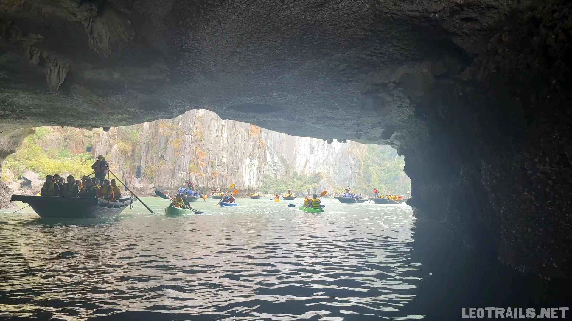 Eye-level view from the dim interior of Luon Cave in Ha Long Bay, Vietnam, looking out through a low limestone archway toward a sunlit emerald lagoon crowded with tourists in small wooden rowboats and colorful kayaks under towering, distant karst cliffs.