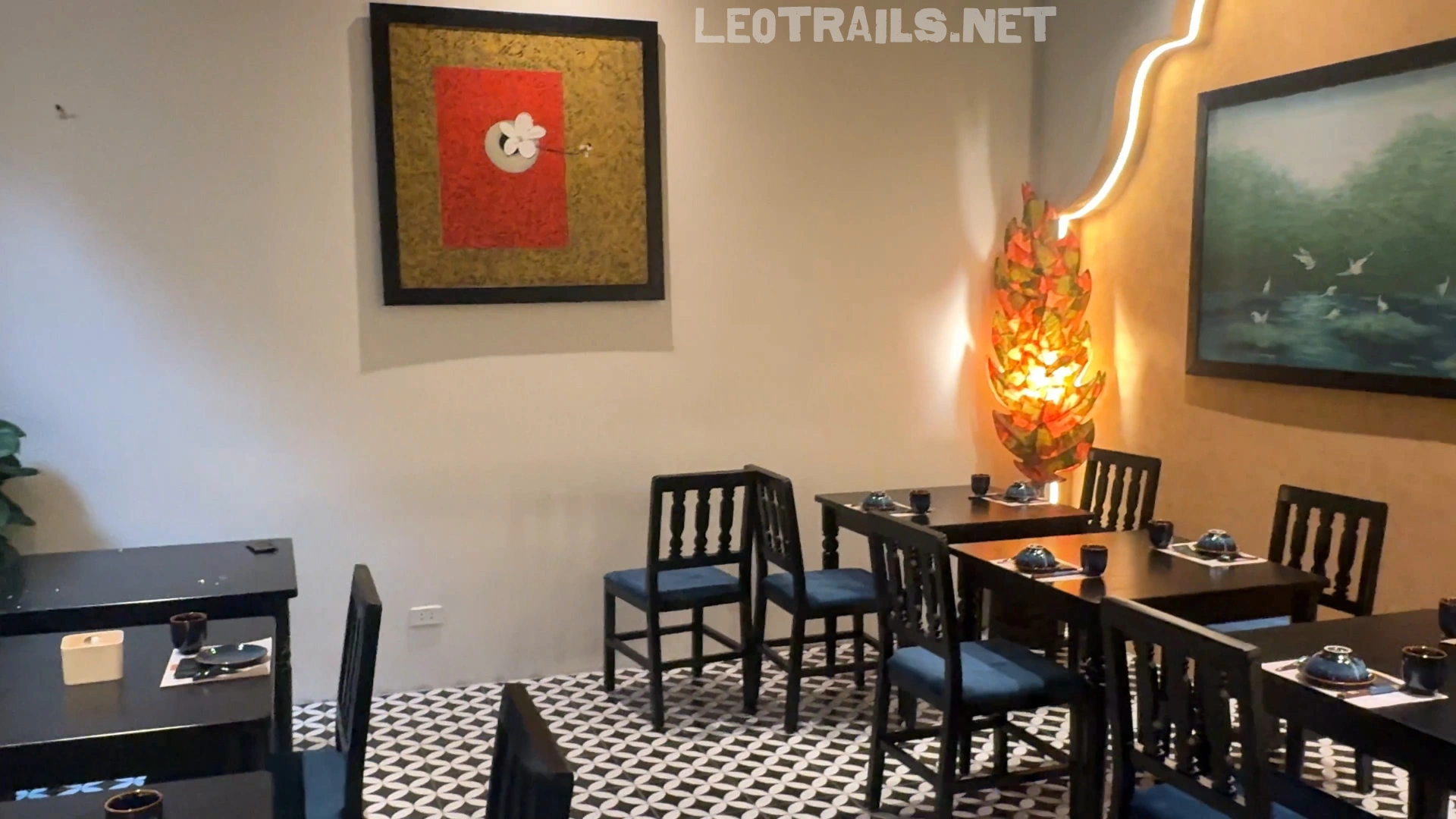 The indoor dining area of Madam Tư Hồng restaurant featuring dark wooden tables and chairs, geometric patterned floor tiles, framed artwork, and a unique leaf-shaped floor lamp.