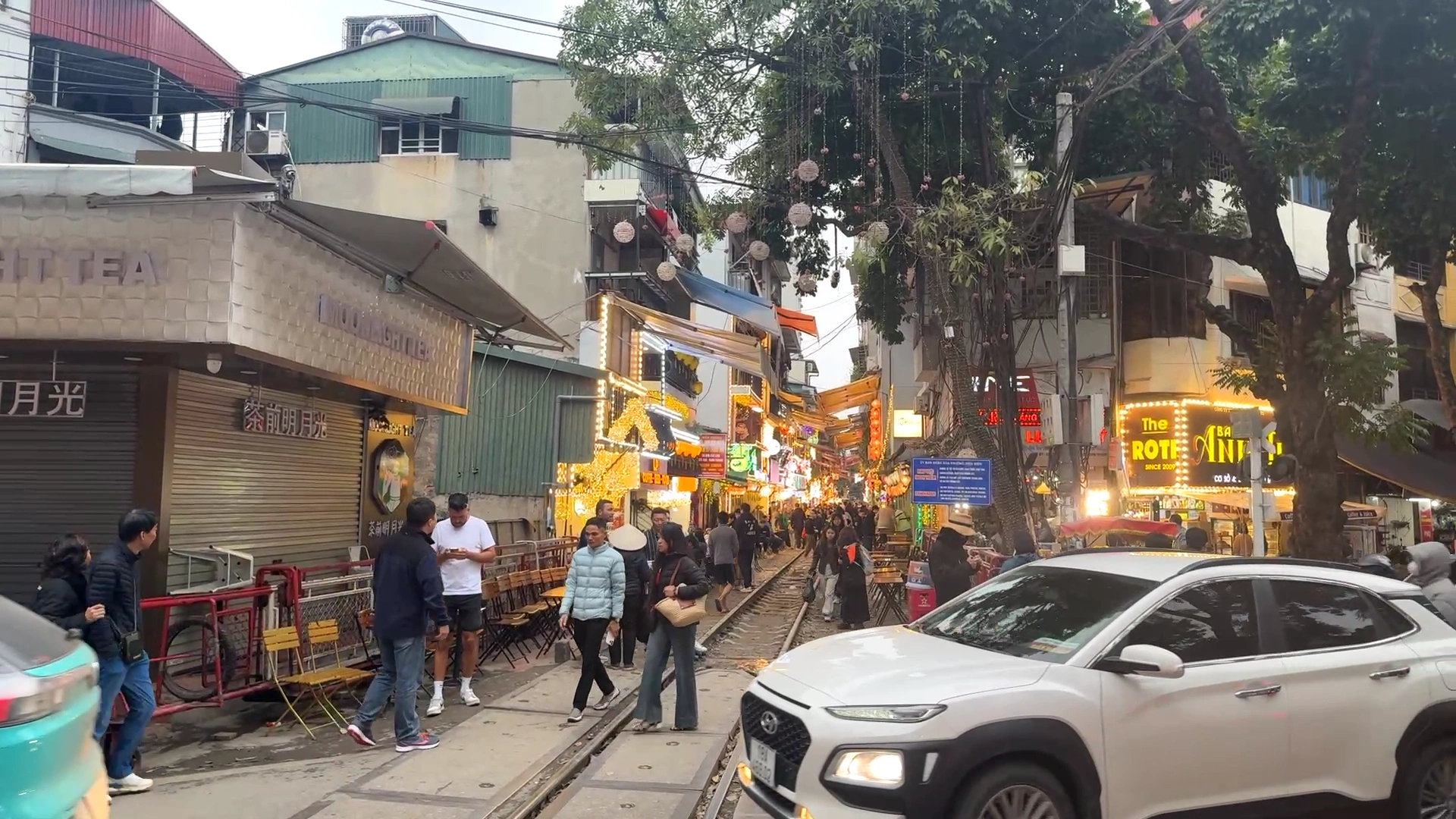 A bustling view of Hanoi Train Street with tourists walking along the railway tracks past lit-up cafes, with a white SUV passing by in the foreground.