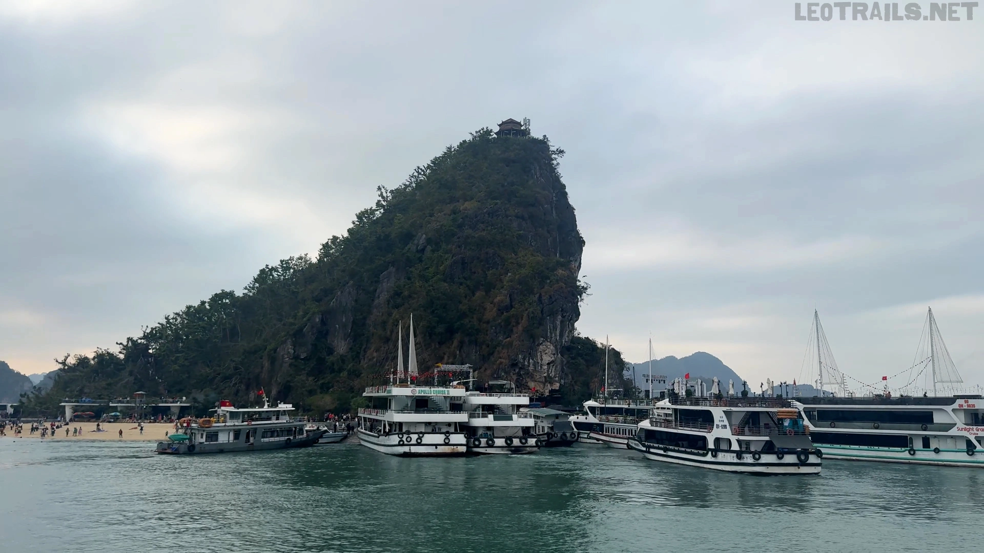 Wide-angle shot of Ti Top Island in Ha Long Bay, Vietnam, featuring a steep, lush green mountain with a traditional viewing pavilion at the peak, multiple white tourist cruise boats docked in the emerald green water, and a sandy crescent beach visible on the left side under an overcast sky.