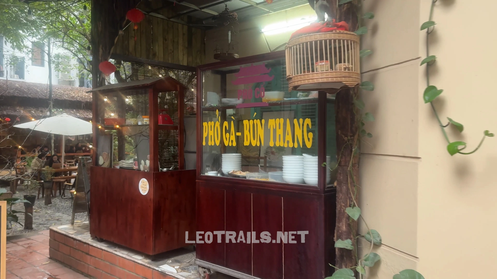 A traditional wooden food cart with a glass display case reading "Phở Gà - Bún Thang" in yellow lettering, situated next to an outdoor restaurant courtyard.