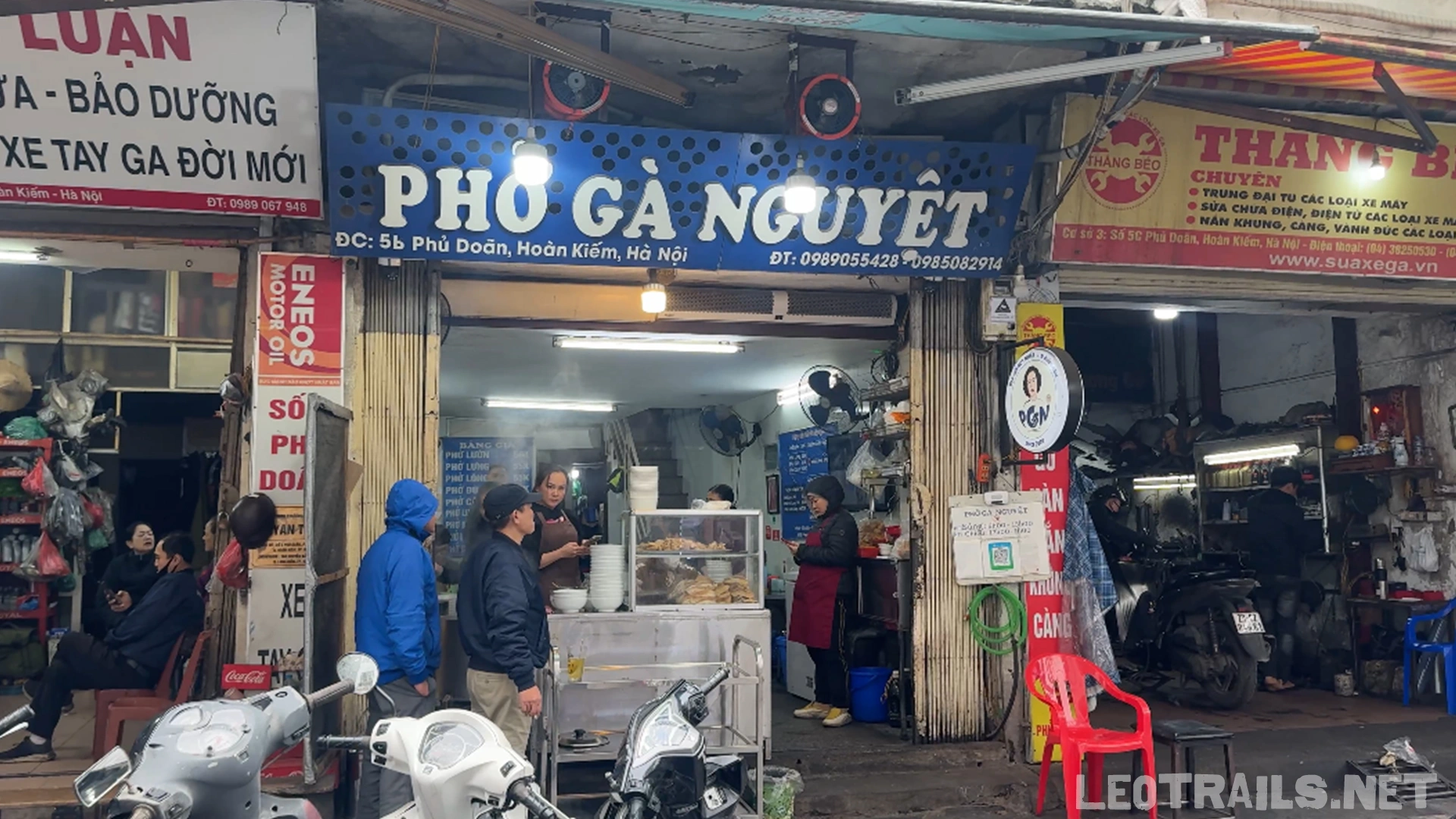 The street-facing storefront of Phở Gà Nguyệt at 5b Phủ Doãn in Hanoi, Vietnam, showing a blue and white sign, customers waiting outside, and motorbikes parked in front.