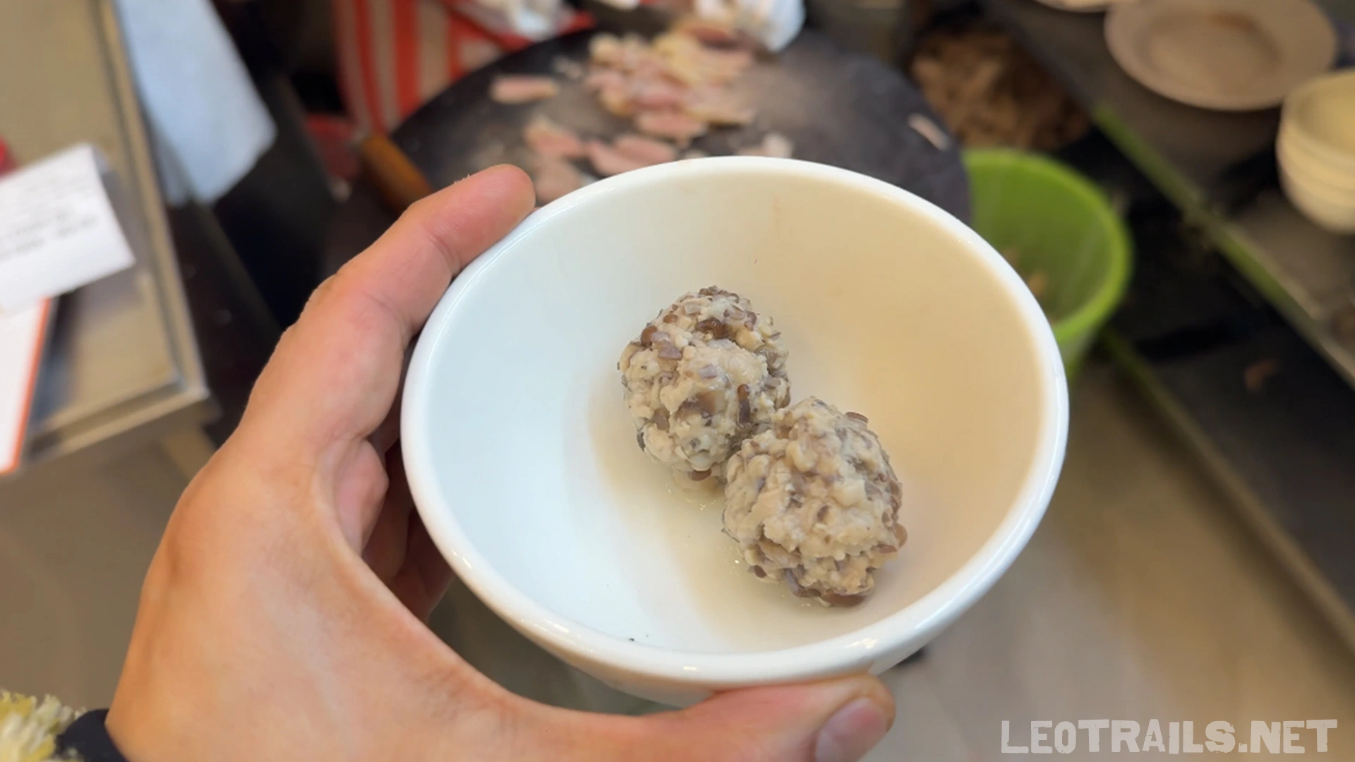 A hand holding a small white ceramic bowl containing two round duck meatball paste portions (mọc ngan) mixed with wood ear mushrooms, ready to be cooked in hot broth.