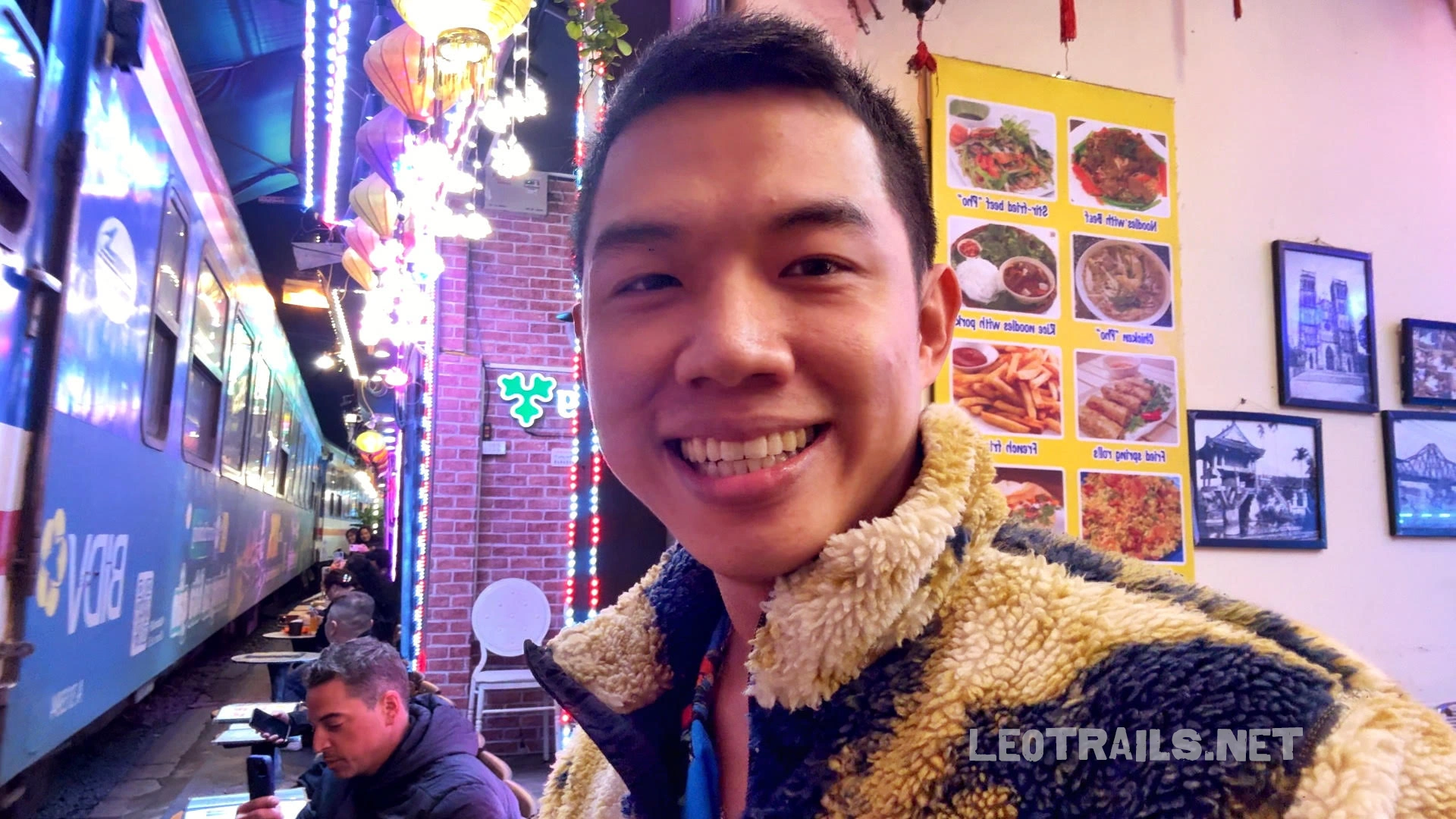 I am taking a selfie at a Hanoi Train Street cafe as a blue train passes closely beside the outdoor seating area at night.