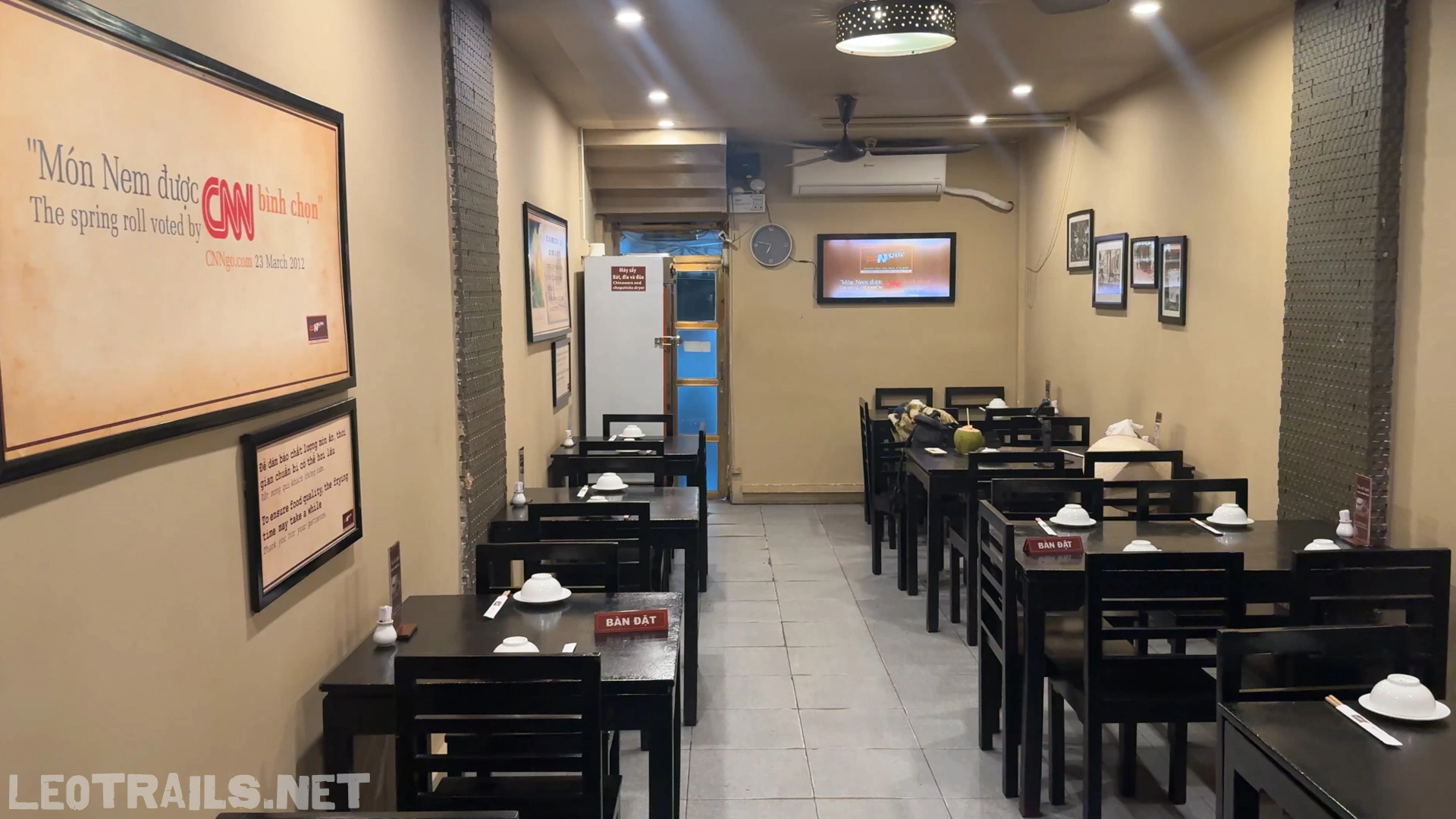 The indoor dining room of Quán Nem restaurant featuring dark wooden tables and chairs, with a framed sign on the left wall highlighting a CNN voting feature for their spring rolls.
