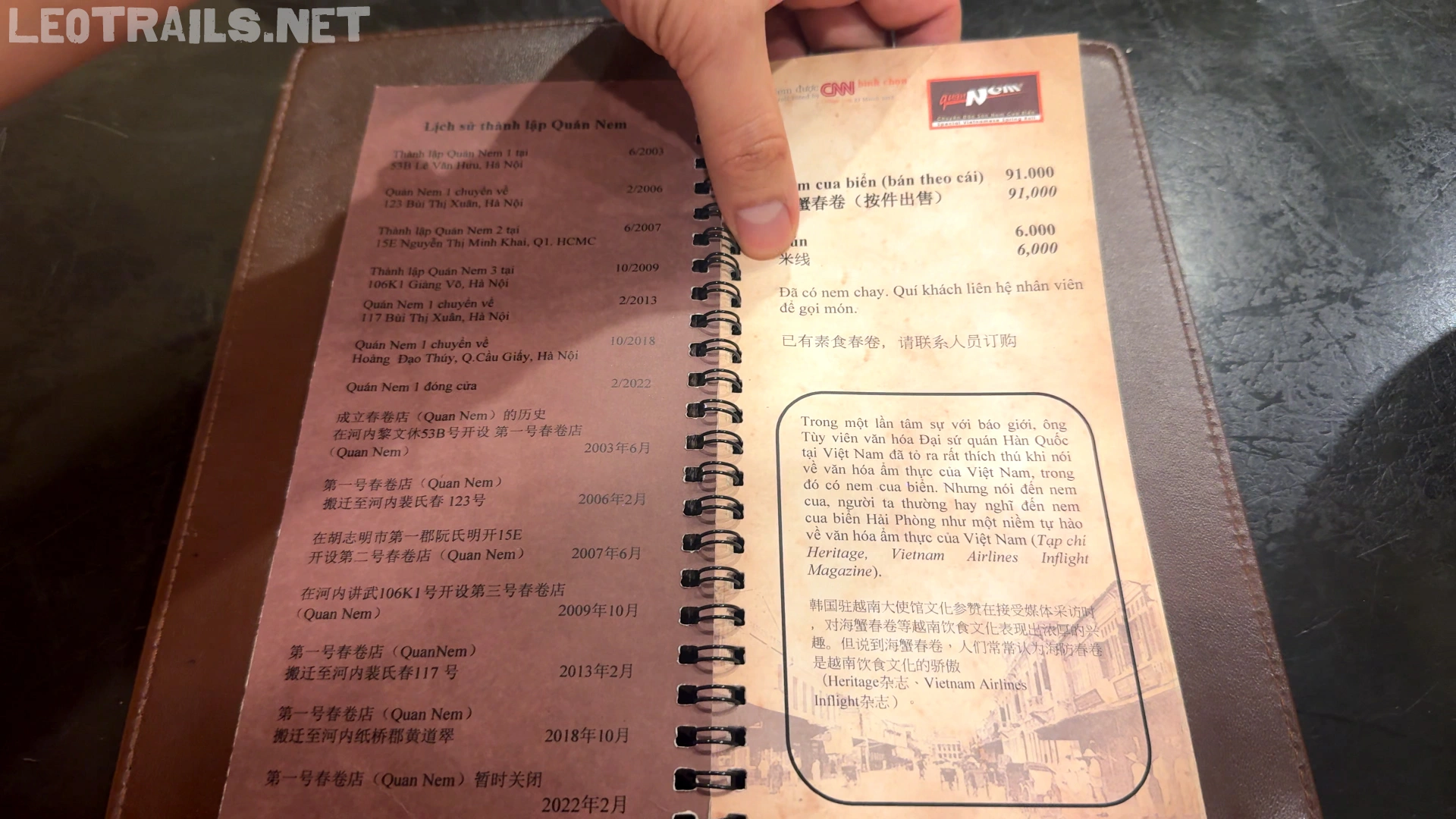 A hand pointing to a spiral-bound menu booklet at Quán Nem restaurant, showing prices for sea crab spring rolls listed at 91,000 VND and a chronological history of the restaurant on the left page.