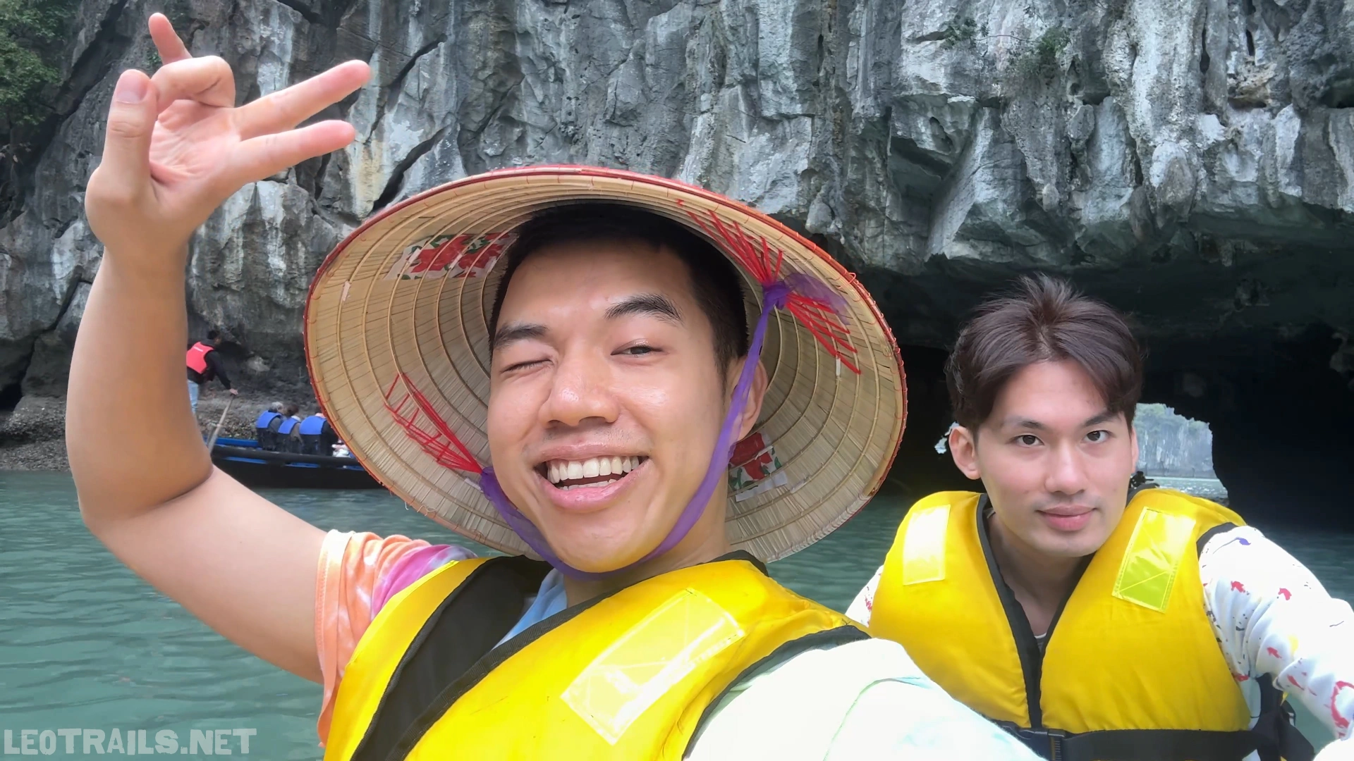 Close-up selfie of two cheerful male tourists in bright yellow life jackets paddling a tandem kayak in Ha Long Bay, Vietnam. The man in the foreground is winking and smiling broadly while wearing a traditional Vietnamese conical straw hat (Non La) and making a hand gesture, with towering limestone karst cliffs in the background.