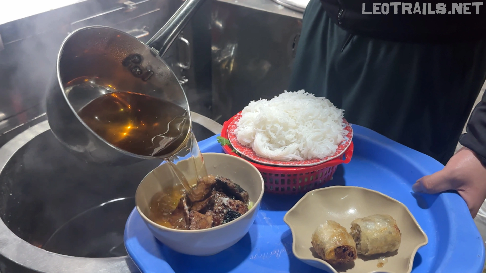 A vendor pouring warm amber fish sauce broth from a metal ladle over grilled pork in a bowl, arranged on a blue plastic tray with fresh rice vermicelli noodles and sliced fried spring rolls