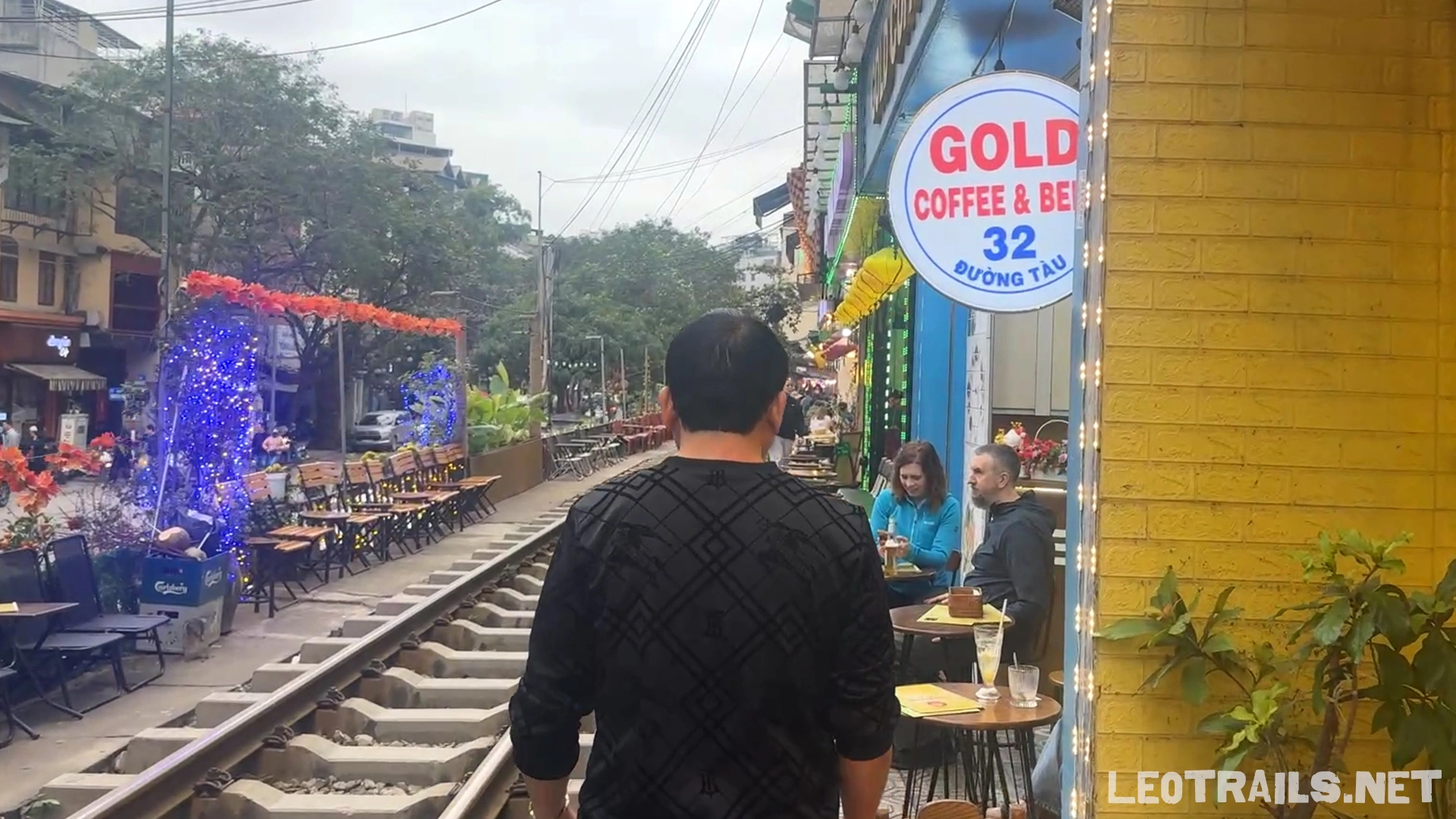 A man in a black pattern shirt seen from behind, walking down the railway tracks past sidewalk cafes, including one with a round blue sign reading Gold Coffee & Beer.