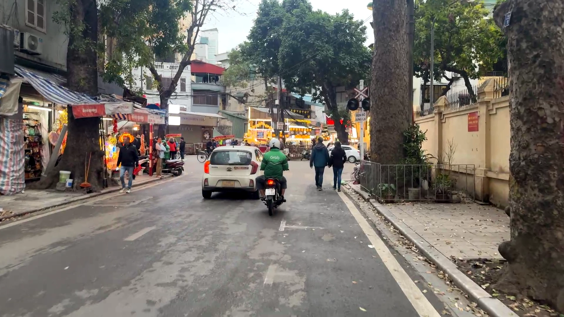 A street view in Hanoi showing a Grab motorbike driver and a white car driving down a road lined with trees and shops toward a busy intersection.