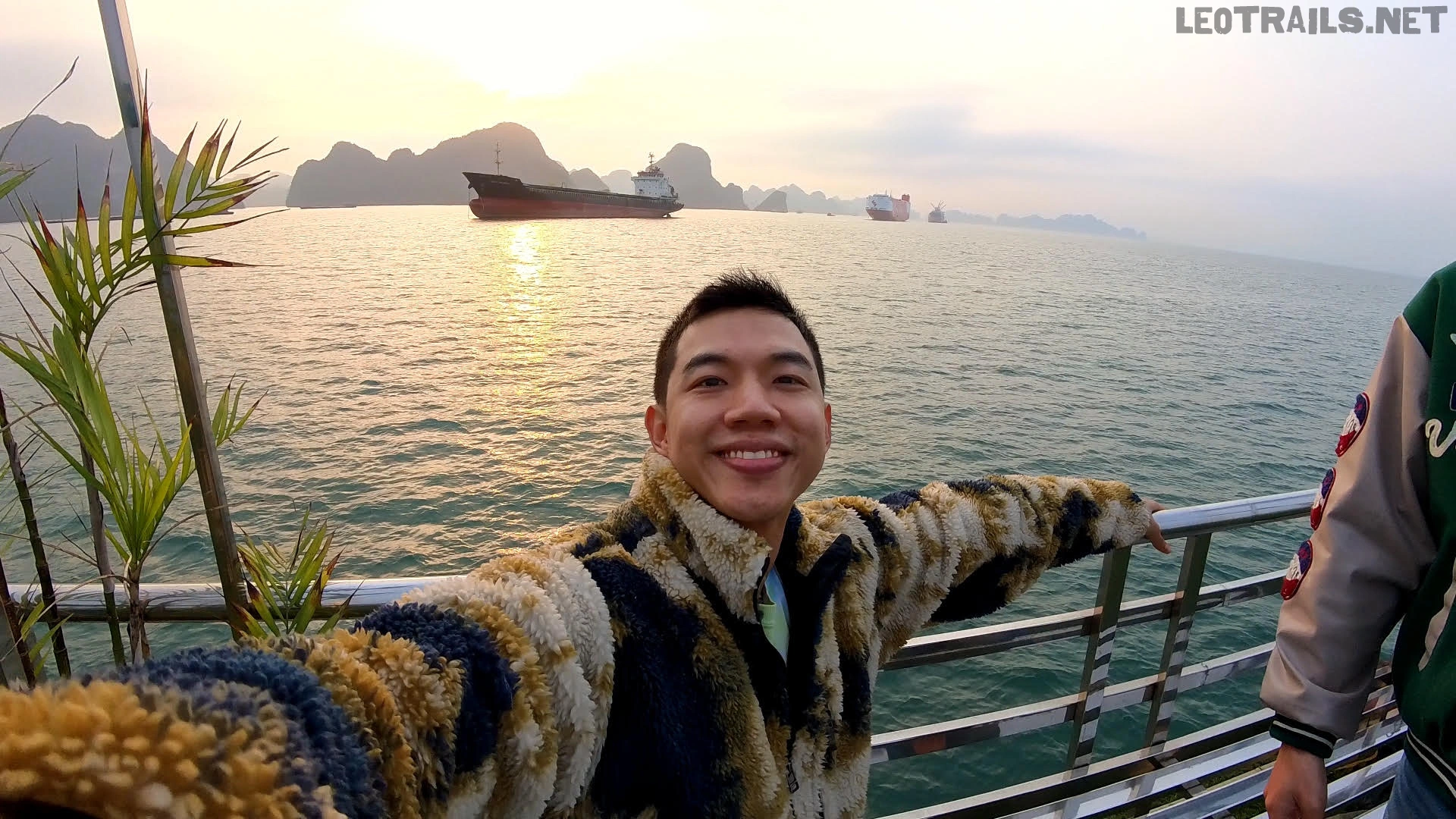 A smiling man taking a selfie on a cruise ship deck with arms outstretched, against a backdrop of a golden sunset, calm sea, and cargo ships near limestone islands.