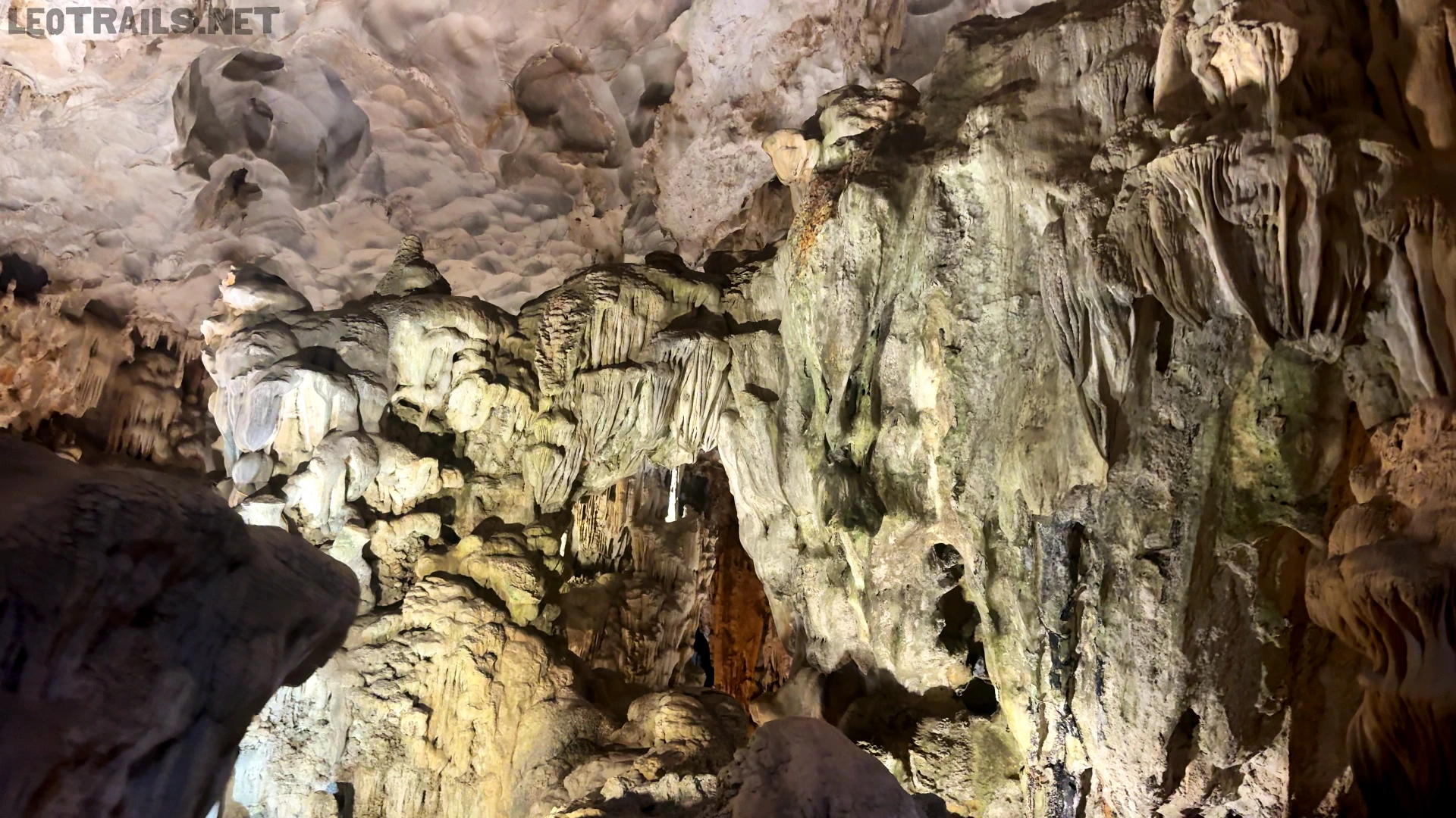 Massive, intricately layered limestone formations and flowing rock textures illuminated inside a large cave chamber.