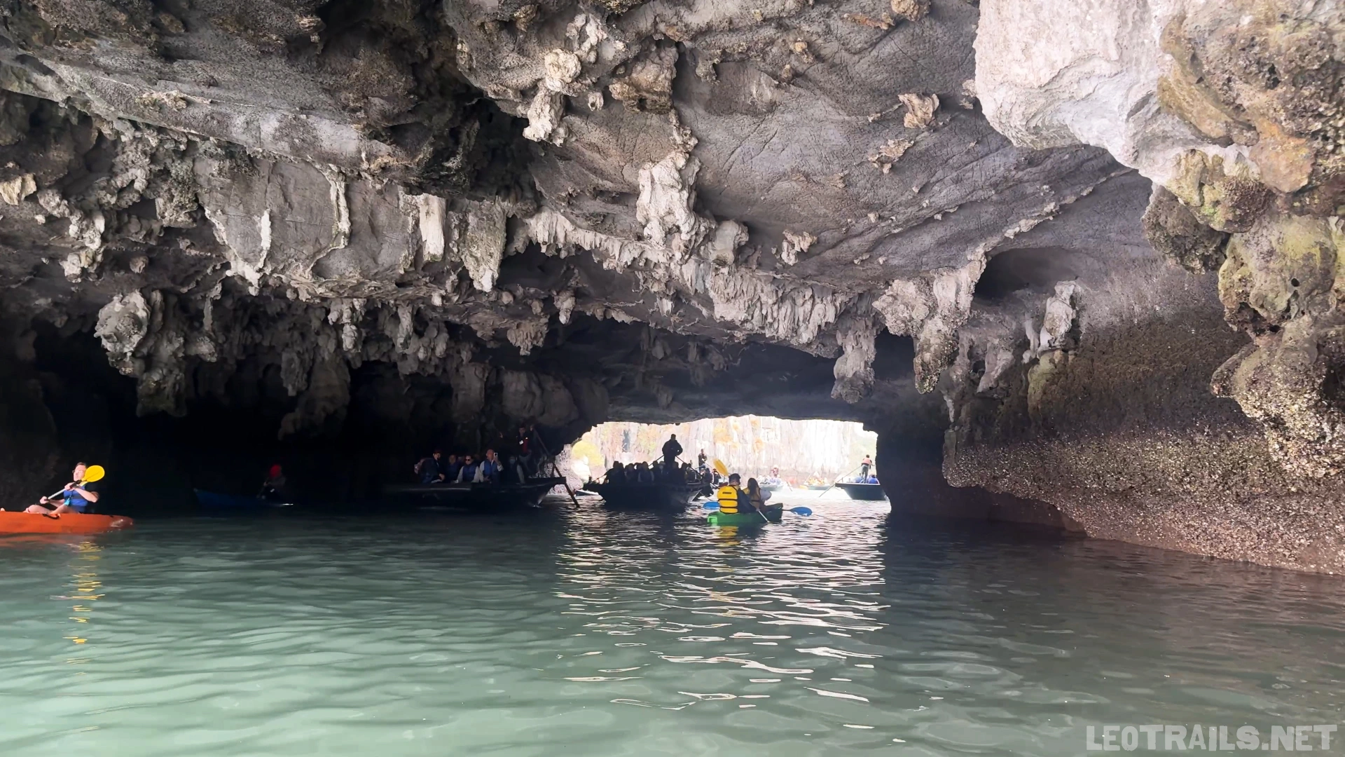 Wide-angle interior view from inside Luon Cave water tunnel in Ha Long Bay, Vietnam. Low-hanging jagged limestone stalactites line the dark cavern ceiling above emerald water, where several tourist rowboats and an orange kayak navigate toward the bright arched exit in the distance.